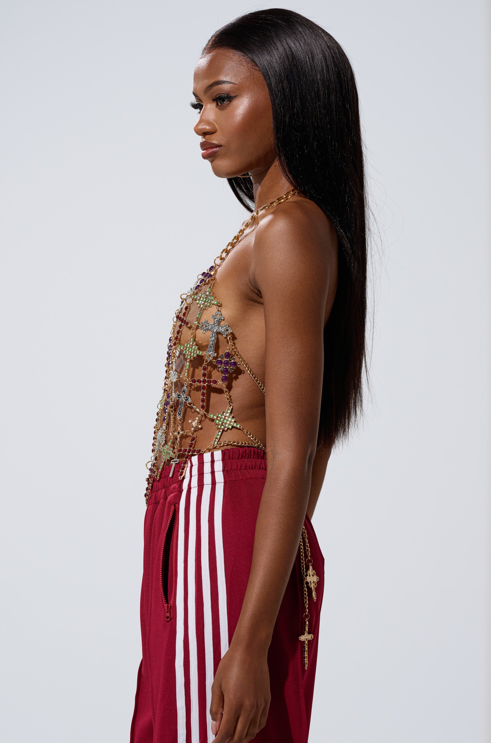 A woman stands in profile wearing the BLESSED BRALETTE and red track pants with white stripes. Her long, straight black hair frames her neutral expression against a plain light background.