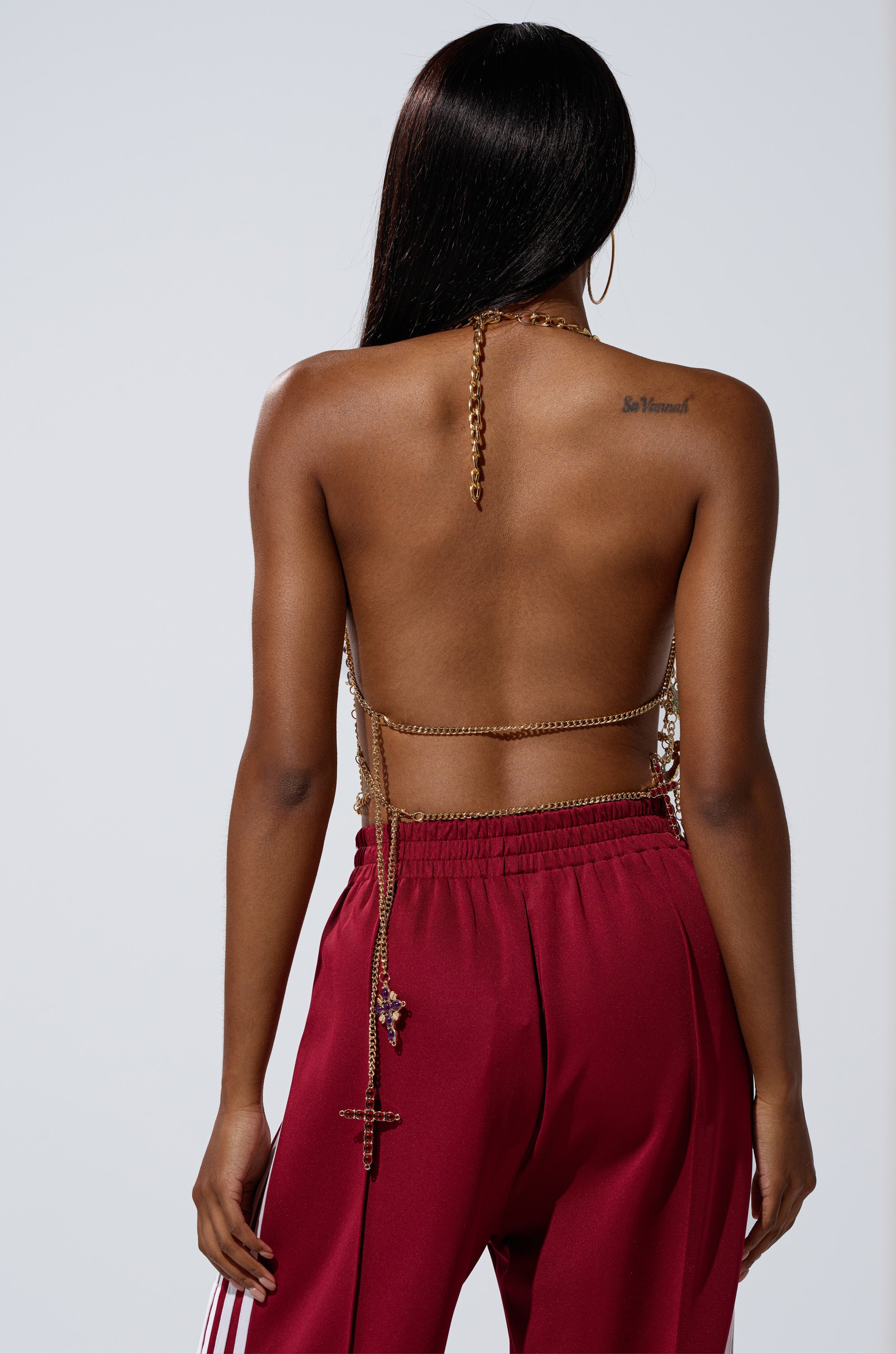 A woman faces away, showcasing her bare back with the BLESSED BRALETTE—a gold chain bralette draped over her shoulders and a tattoo on her right shoulder—paired with maroon pants featuring white side stripes.