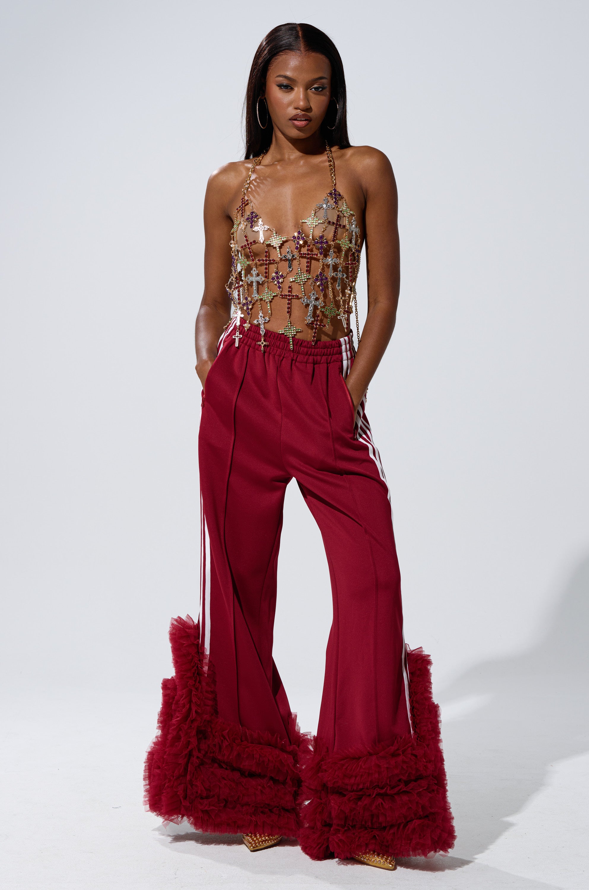 A woman poses confidently in the BLESSED BRALETTE and high-waisted maroon pants with ruffled flares, styled with hoop earrings and pointed shoes, standing out against a plain background.