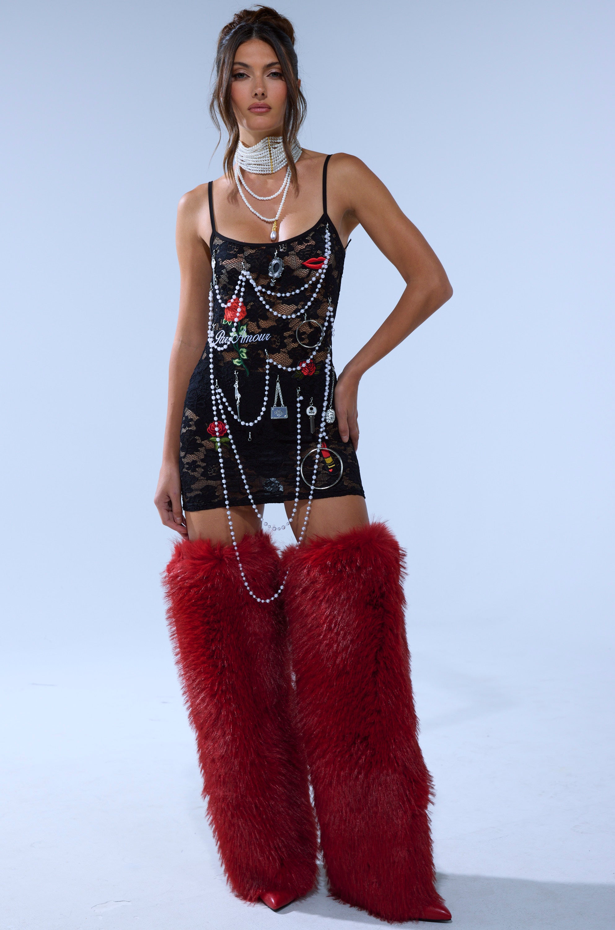 A woman in the FEELING CRAZY SEAMLESS JUNK MINI DRESS, styled with layered pearl chains and red faux fur knee-high boots, stands against a plain light background.