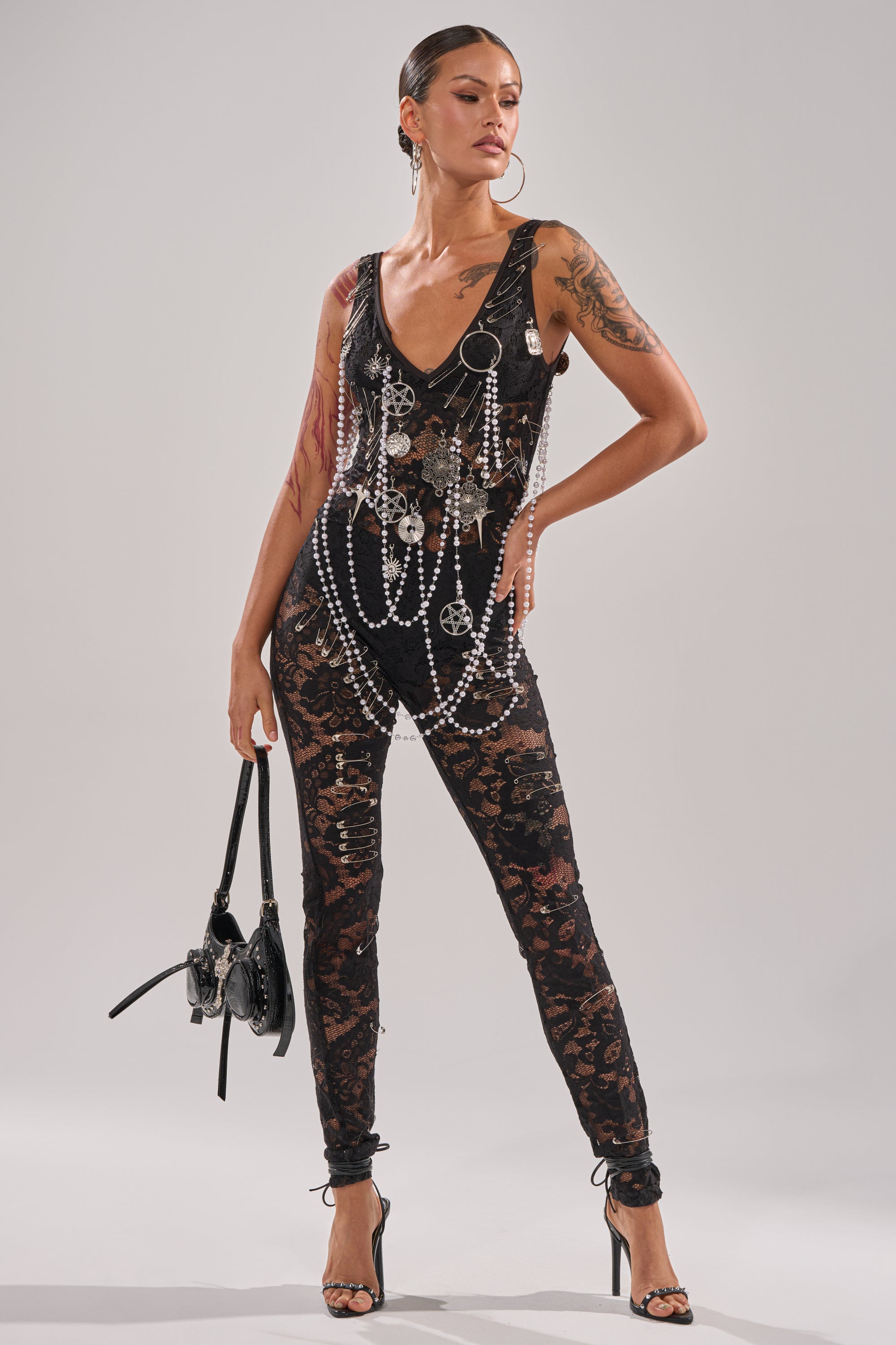 A person stands confidently against a plain light background wearing the BABY I AM THE PARTY LACE JUMPSUIT— a sheer floral lace piece styled with layered chains, pearls, hoop earrings, high heels, and a black handbag.