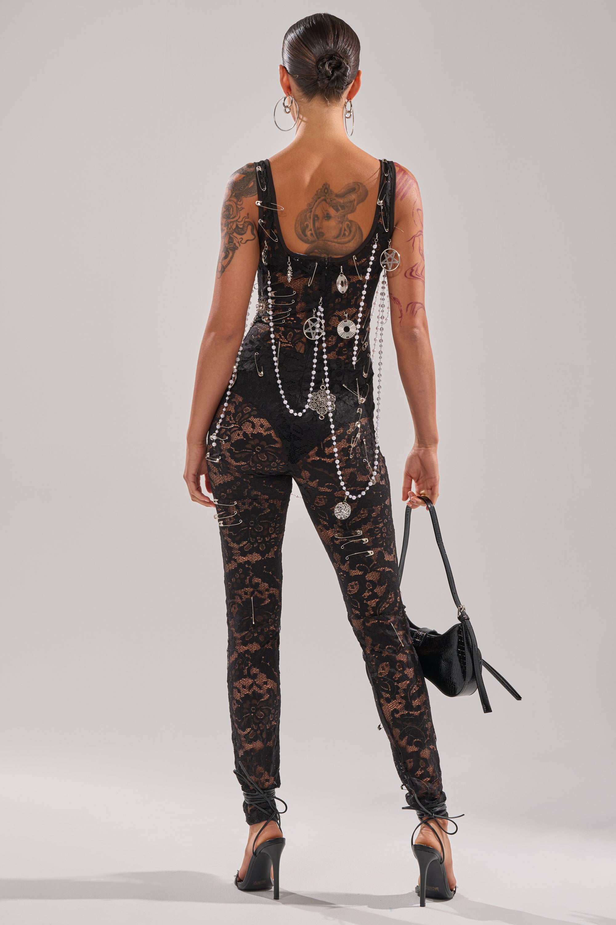 Clad in the BABY I AM THE PARTY LACE JUMPSUIT with chains and pearls, a person faces away to reveal their tattoos, holding a small black handbag. Their hair is in a sleek bun and they stand tall in high heels.