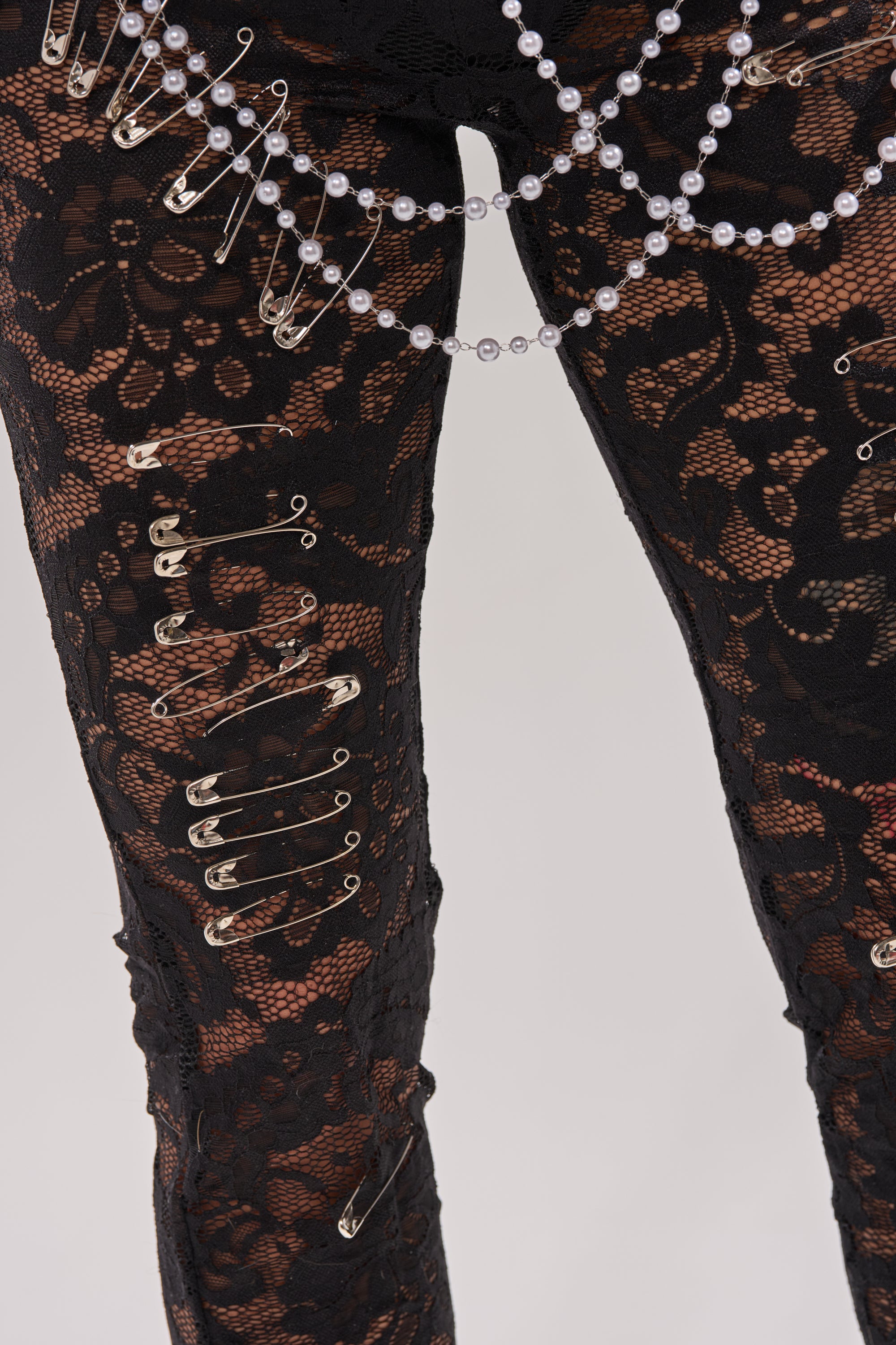 Model in the BABY I AM THE PARTY LACE JUMPSUIT—black, curve-hugging sheer floral lace with silver safety pins and pearls draped at the hip, standing out against a plain light background.