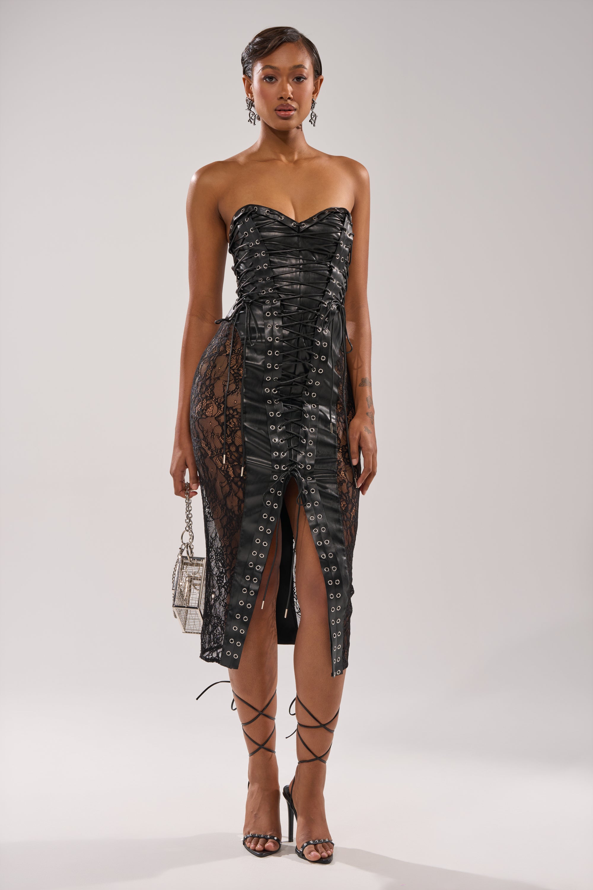 A woman wears the PUNK ROCK LEATHER MIDI DRESS—a strapless black midi with grommet and lace-up details, front slit, and black strappy heels—holding a silver handbag against a plain gray background.