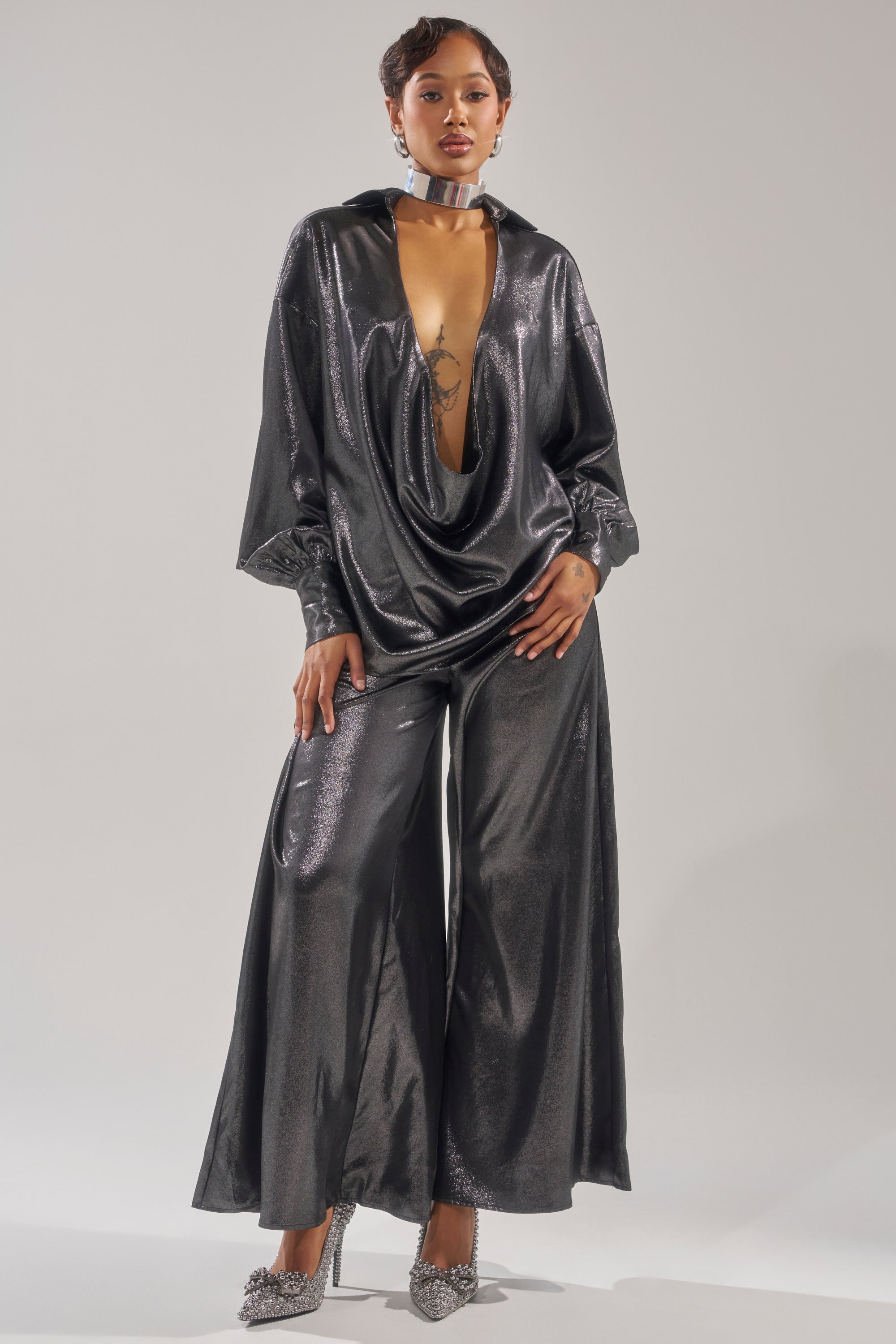 A woman models the MELINIA METALLIC WIDE LEG PANT, featuring glossy metallic fabric and a high-rise fit. She pairs it with a deep cowl neckline top, silver choker, and sparkling heels against a light gray background. 0