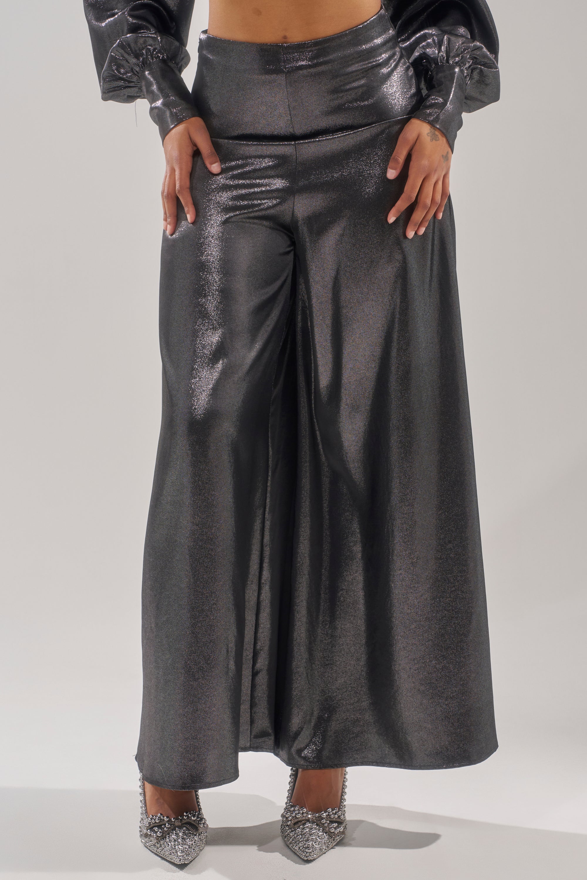 Wearing the MELINIA METALLIC WIDE LEG PANT with a matching long-sleeve top and glittery pointed heels, a person poses with hands on the waistband; the glossy metallic fabric pops against a plain, light background. 2