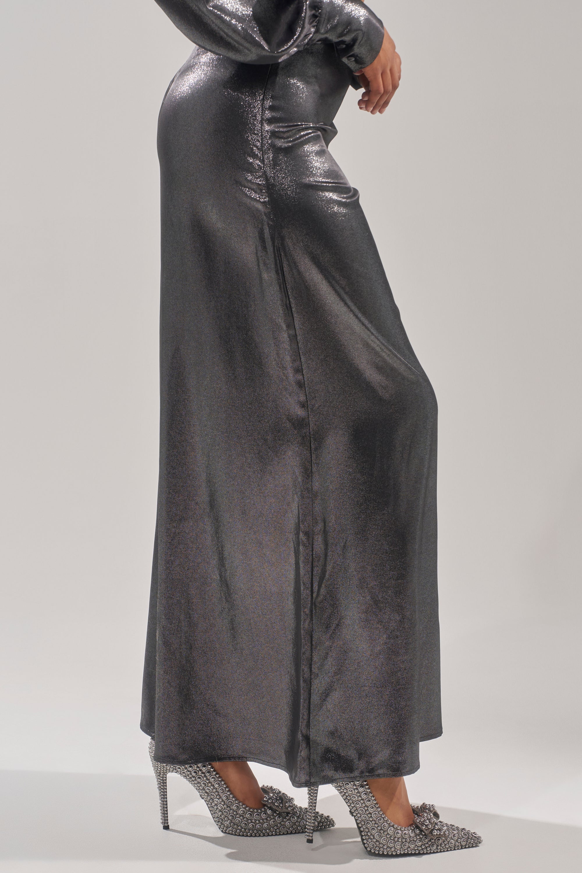 A person wears MELINIA METALLIC WIDE LEG PANT, made from glossy metallic silver fabric, paired with silver embellished high heels, standing against a plain light background. 3