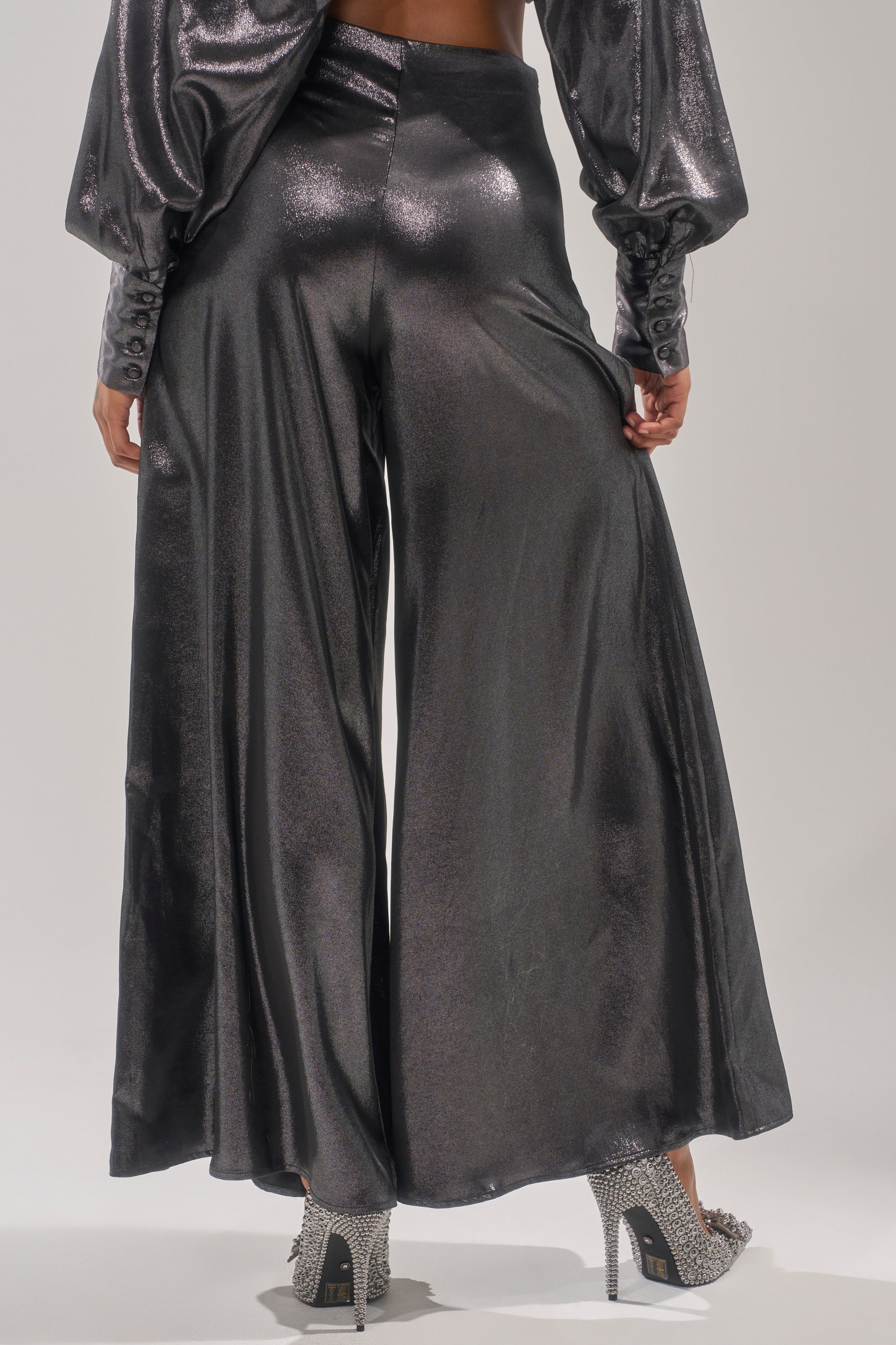 A person is seen from the back wearing the MELINIA METALLIC WIDE LEG PANT with a matching long-sleeve top, holding one side of the pants. The outfit is finished with high-heeled, silver glittery shoes. 4