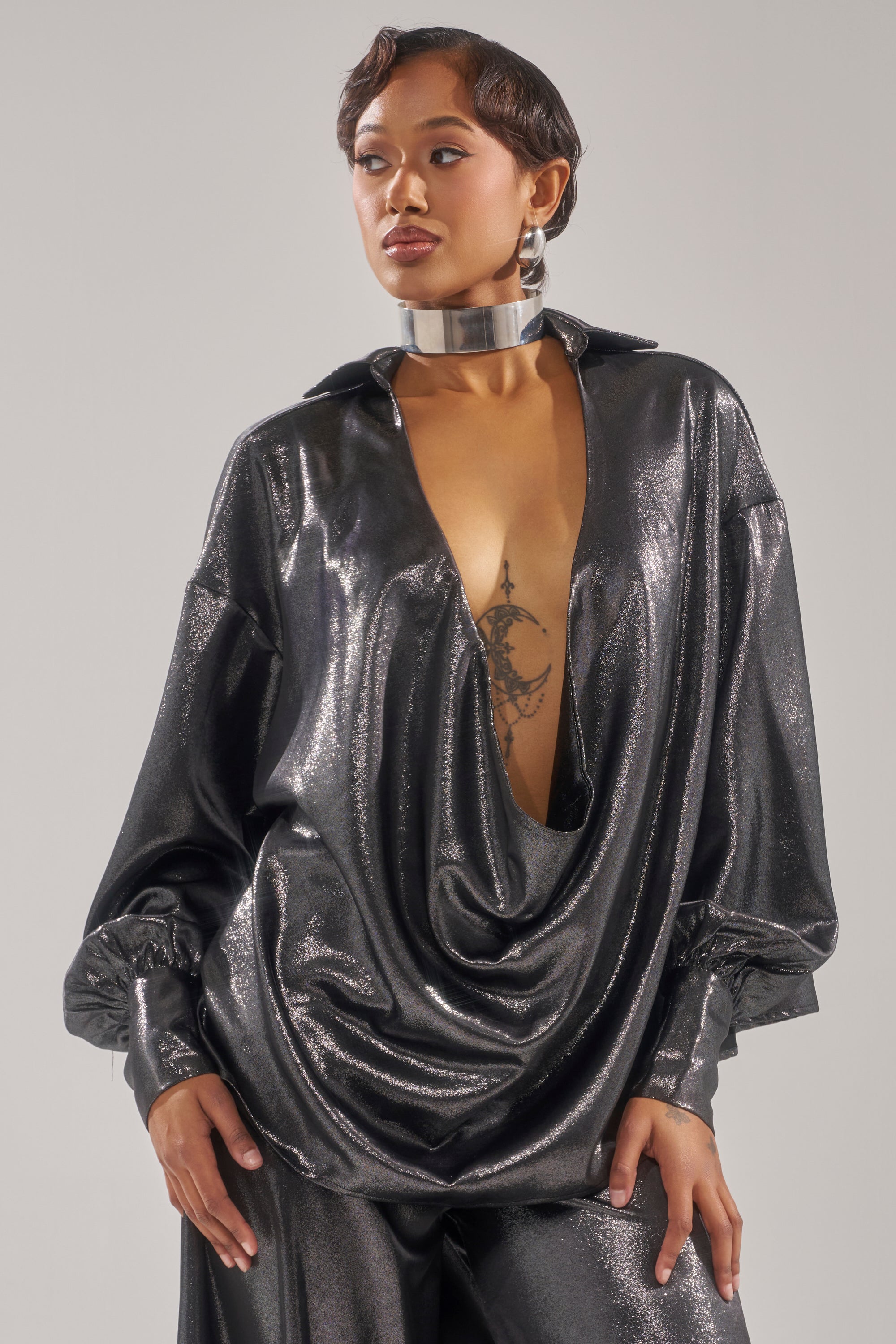 A woman models the MELINIA METALLIC DRAPE BLOUSE with a deep cowl neckline and matching wide-leg pants, accessorized with a silver choker, hoop earrings, and styled hair that highlights her chest tattoo for bold flair.