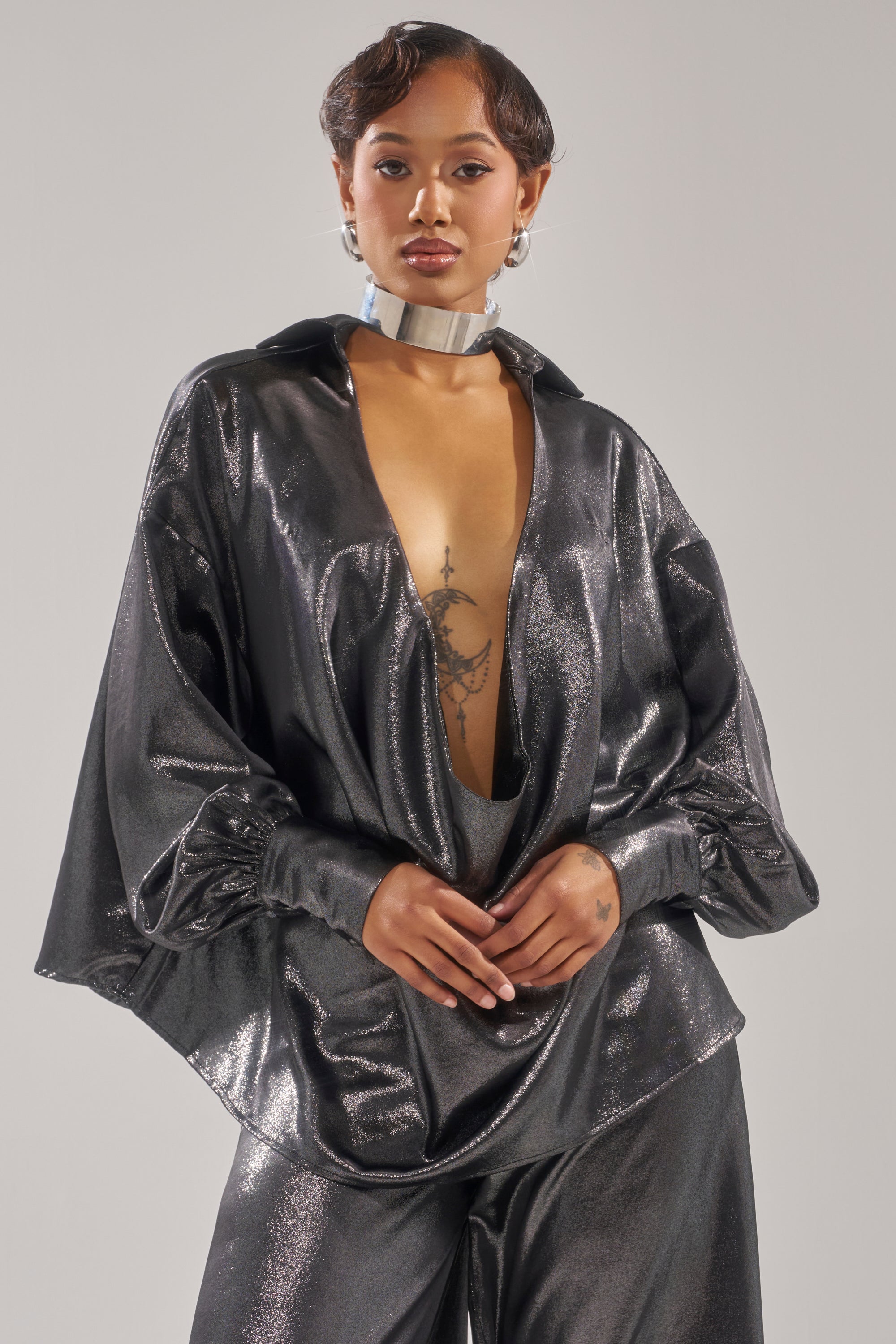 A woman models the MELINIA METALLIC DRAPE BLOUSE with a deep plunging neckline and matching pants, styled with a silver choker necklace and earrings, posing confidently against a plain background.