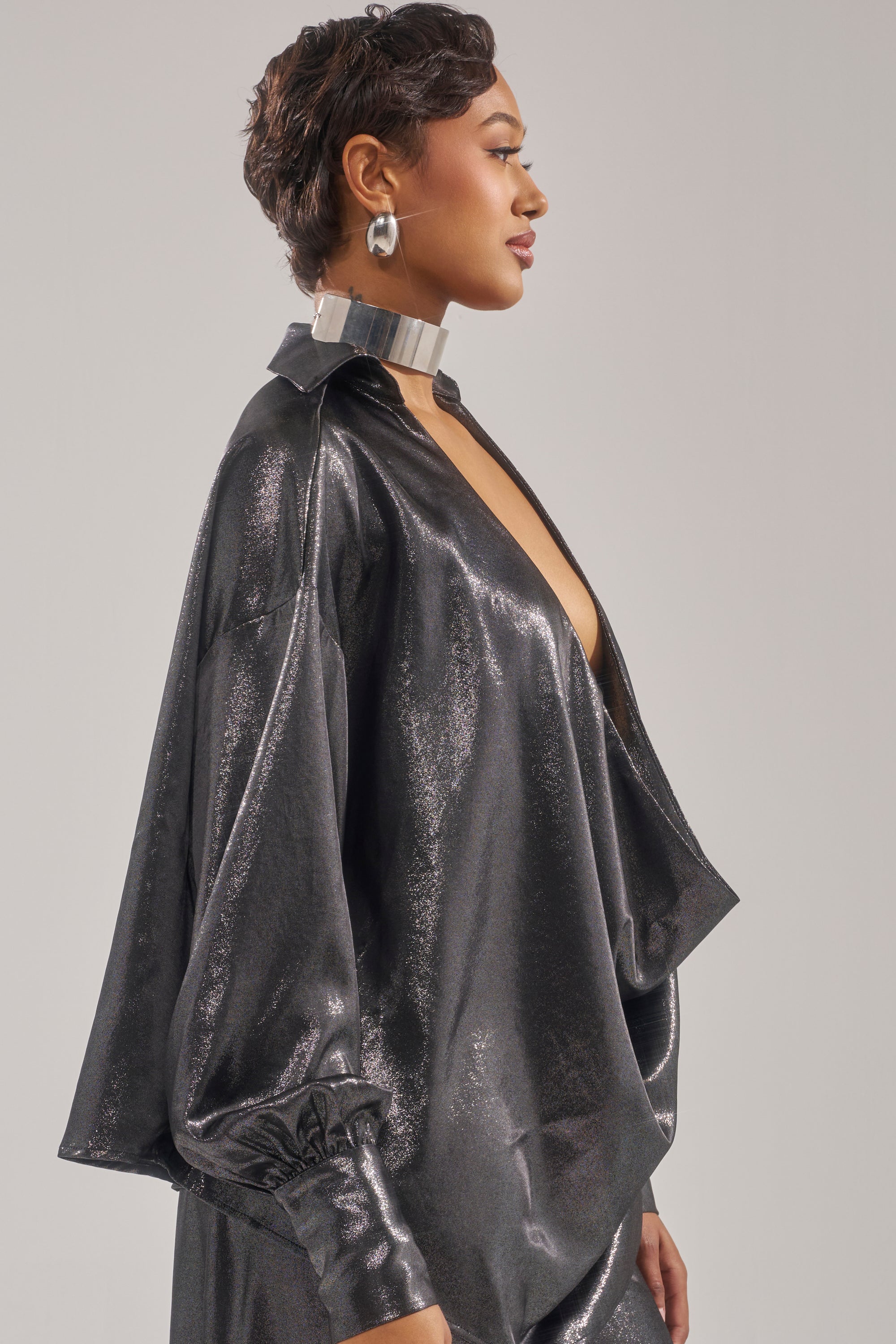 A person in profile wears the MELINIA METALLIC DRAPE BLOUSE in shiny black with voluminous sleeves, paired with a metallic choker and large round earrings, standing against a plain light gray background.