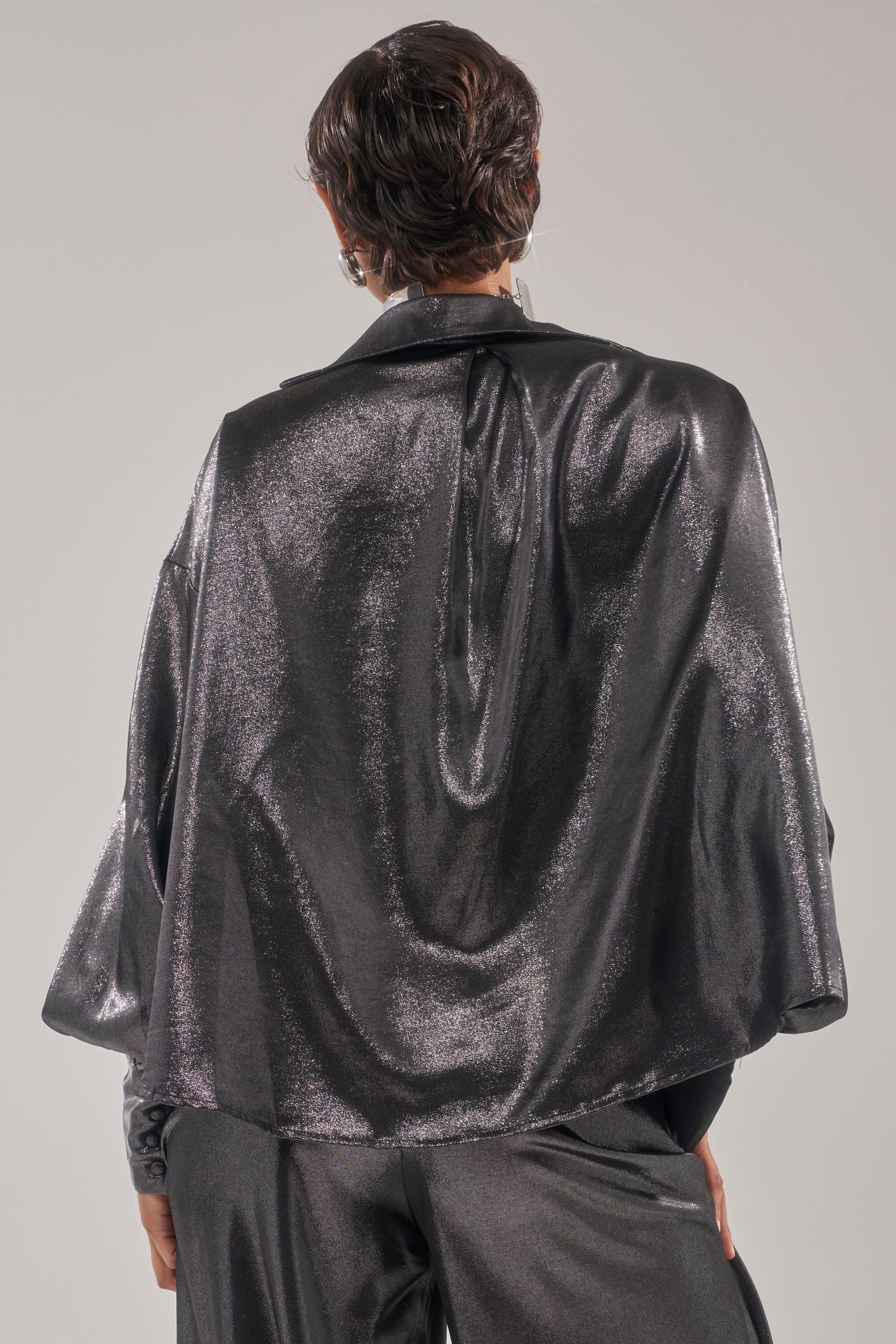 A person with short, dark hair is seen from behind wearing a MELINIA METALLIC DRAPE BLOUSE and matching pants, standing against a plain light gray background.