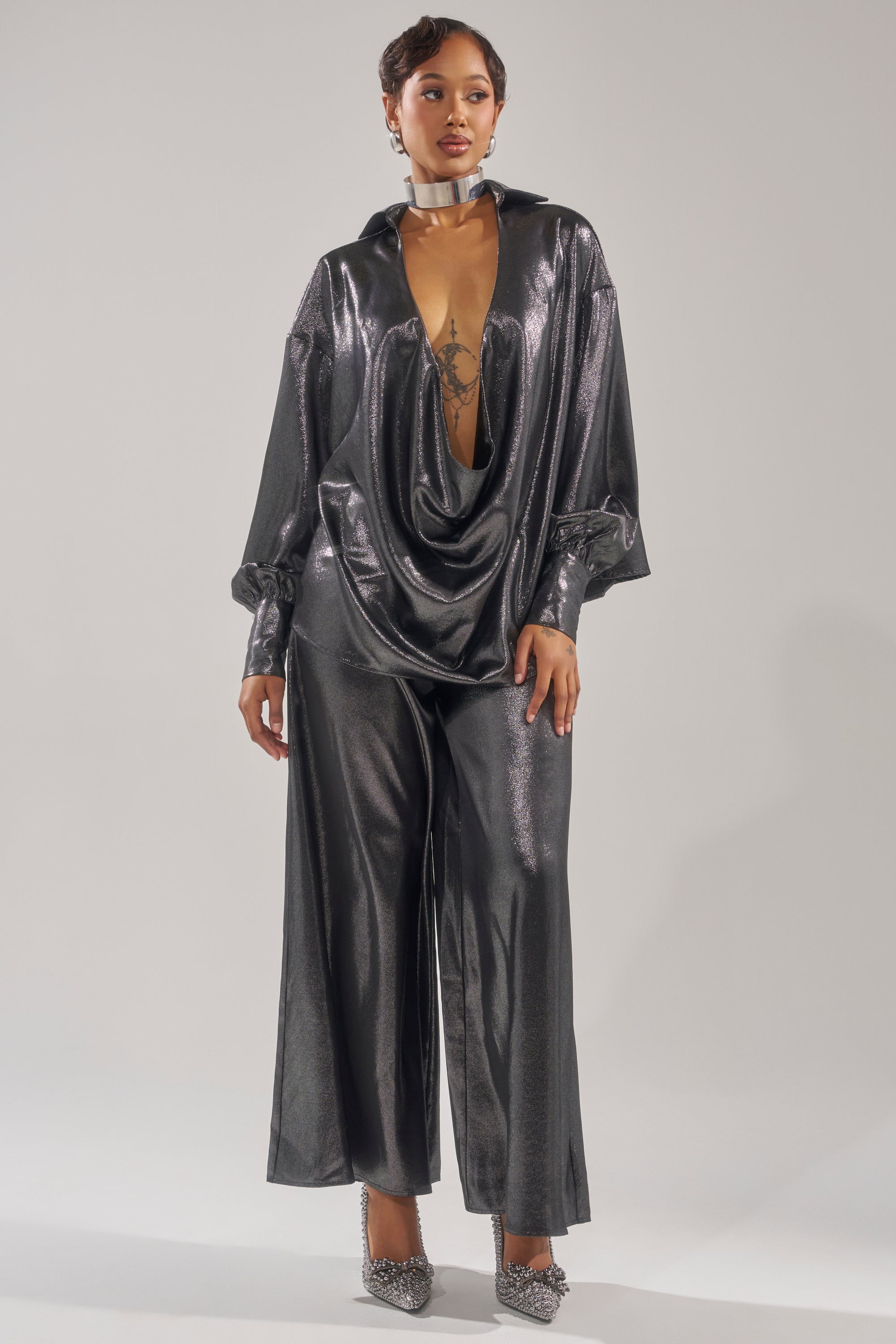 A woman models the MELINIA METALLIC DRAPE BLOUSE with a deep plunging neckline and pairs it with wide-leg pants, a silver choker, and embellished heels, standing against a plain light background.