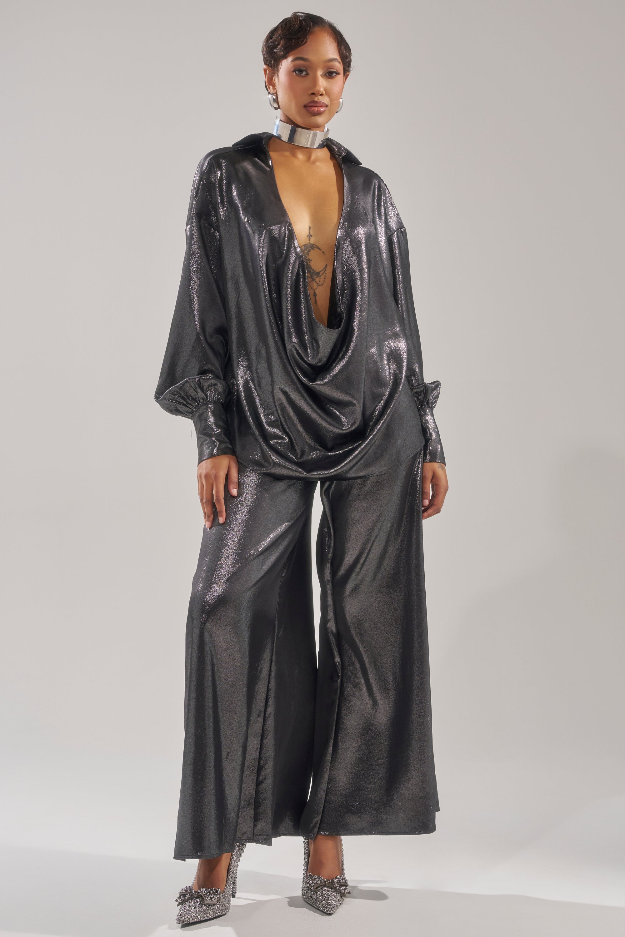 A woman stands confidently against a plain background wearing the MELINIA METALLIC DRAPE BLOUSE paired with wide-leg matching pants, sparkly heels, and a silver choker necklace.