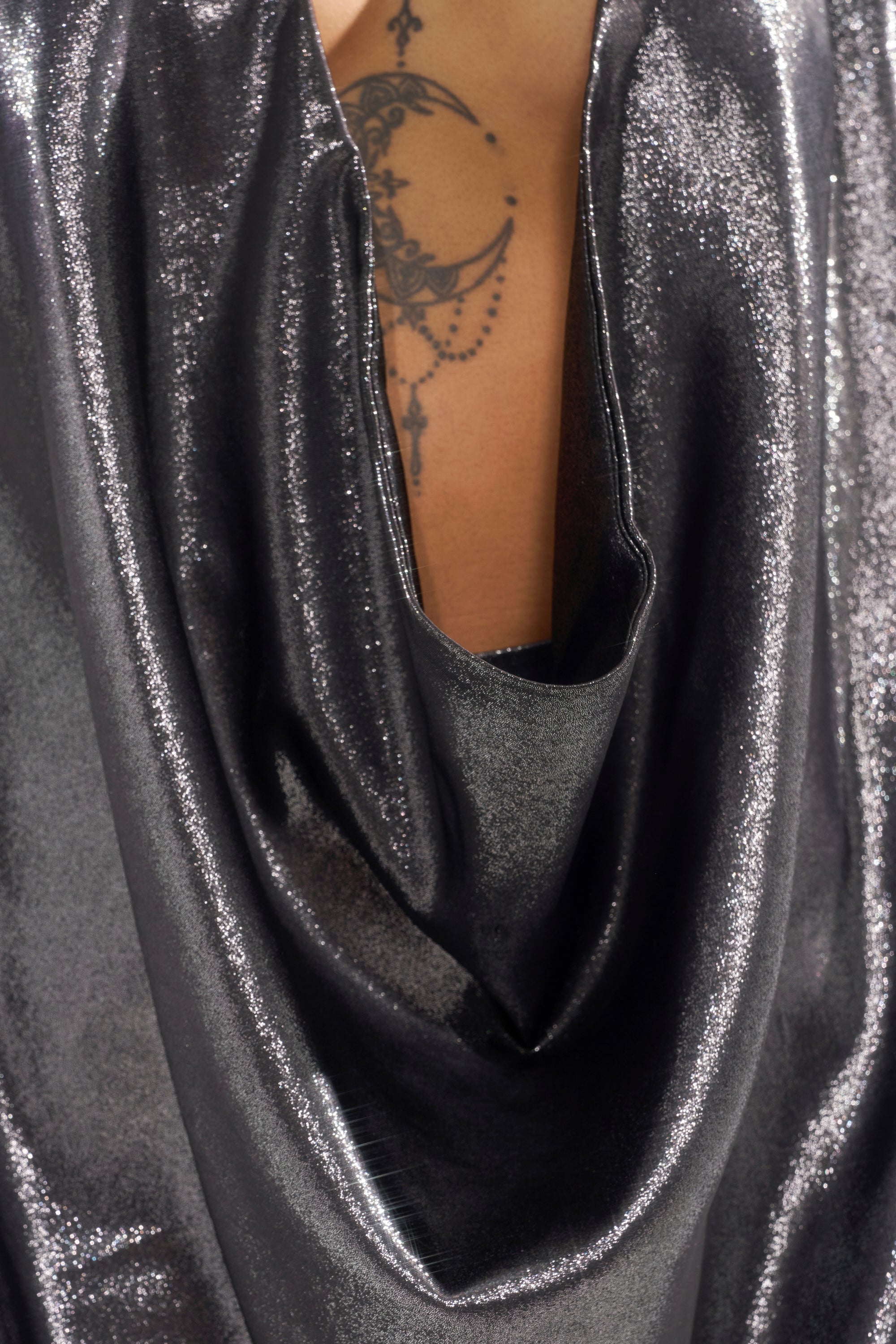 Close-up of a person wearing the MELINIA METALLIC DRAPE BLOUSE, with the shiny fabric draped to reveal part of their chest and a crescent moon tattoo decorated with dots and lines.