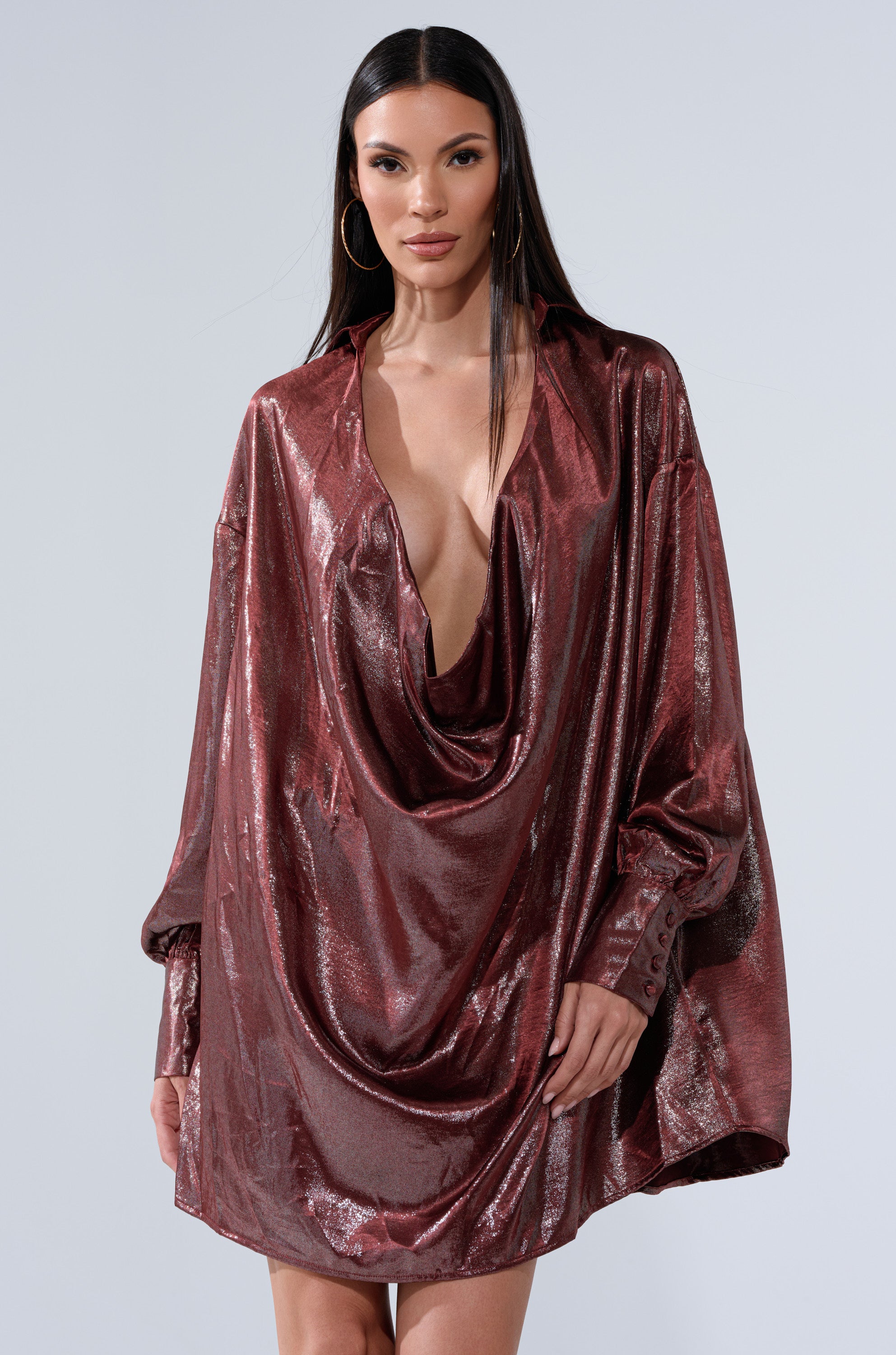 A woman wears the LENNON PLUNGING SATIN SHIRT DRESS—an oversized, metallic burgundy piece with long sleeves and a deep cowl neckline—standing against a plain light gray background.