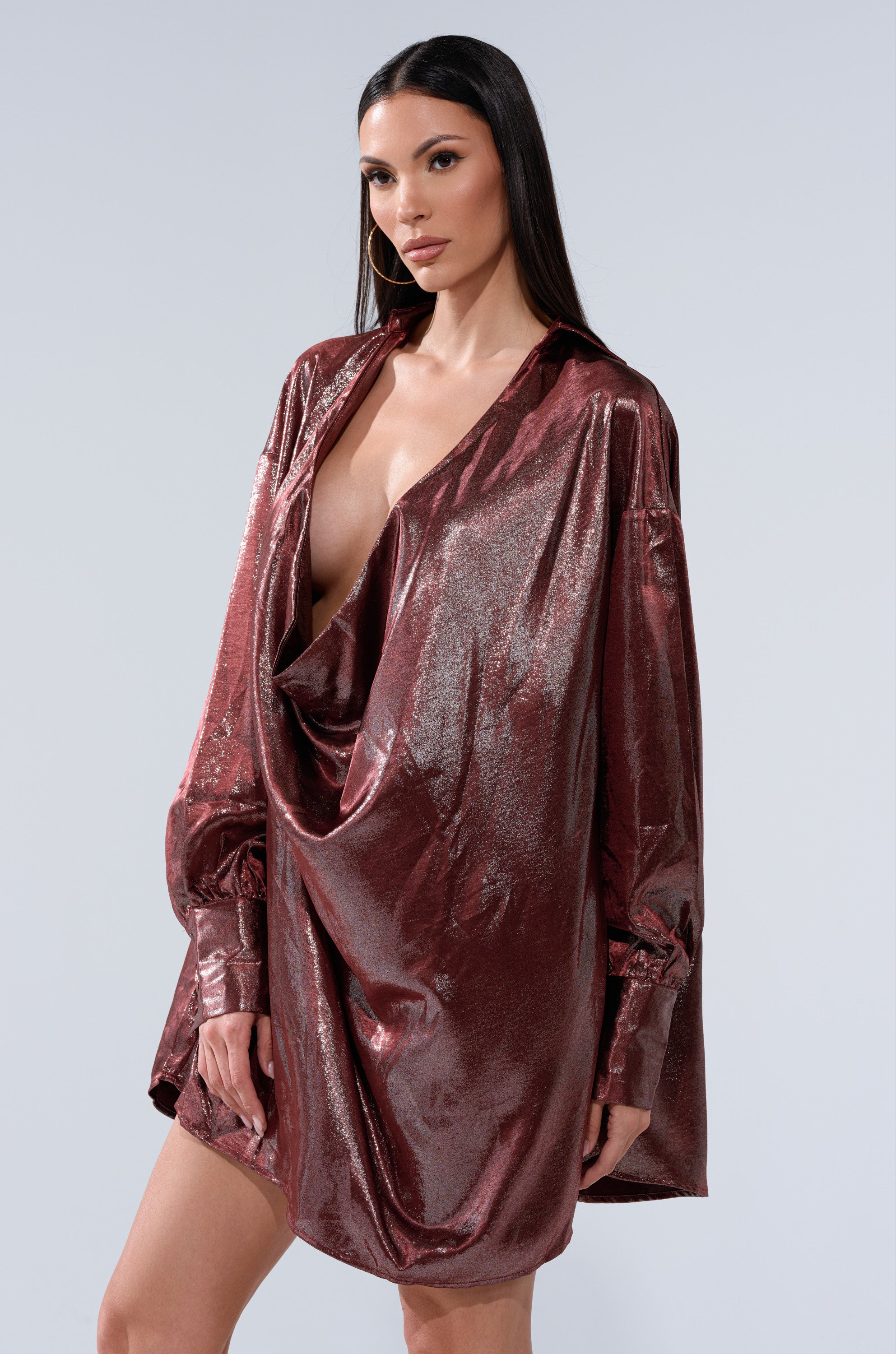A woman stands against a plain light gray background, wearing the LENNON PLUNGING SATIN SHIRT DRESS—a shiny, loose-fitting metallic maroon dress with long sleeves and a deep cowl neckline.