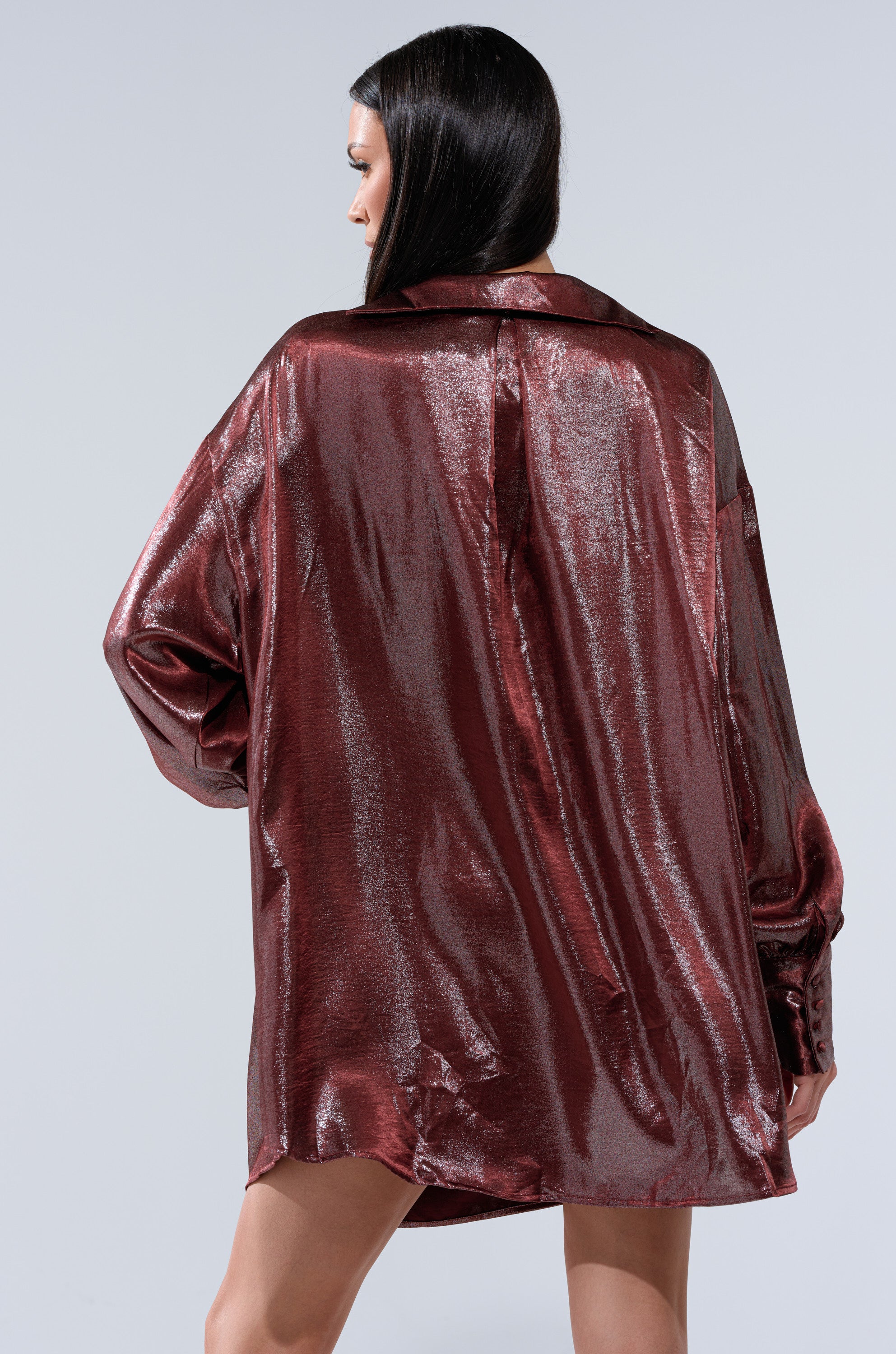 A woman with straight dark hair stands with her back to the camera, wearing the LENNON PLUNGING SATIN SHIRT DRESS in metallic burgundy with long sleeves, set against a plain light gray background.