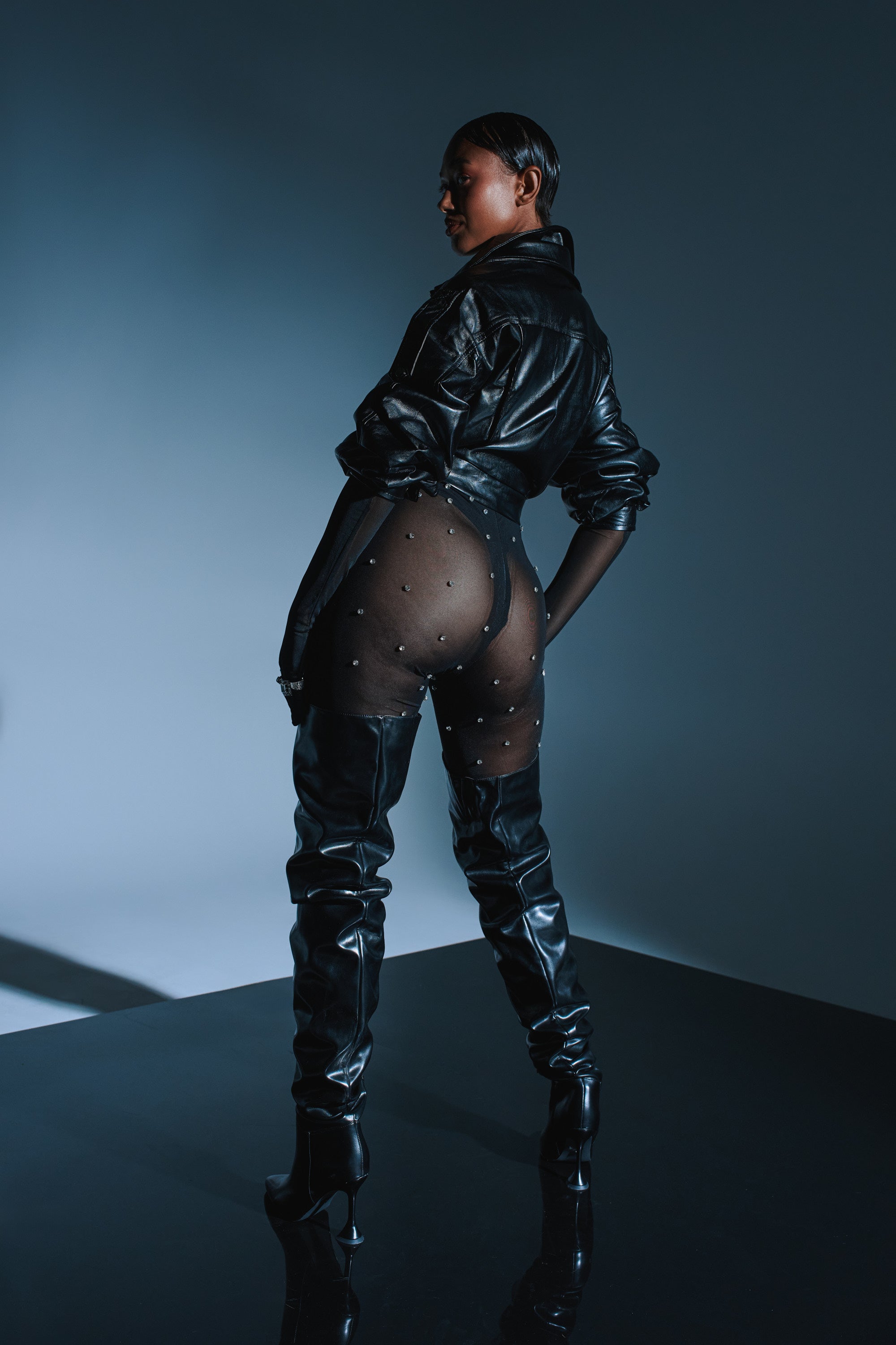 A person stands confidently on a reflective surface in the ALEX RHINESTONE MESH JUMPSUIT, paired with thigh-high black boots and a leather jacket. The dramatic background features soft, directional lighting.