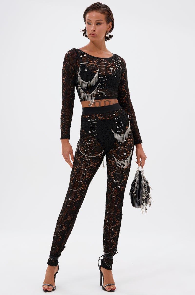 A woman stands confidently against a white background, wearing the TRY ME LACE LEGGING as part of a fitted black lace two-piece with silver chain details, holding a small black handbag and finishing the look with black high-heeled sandals.
