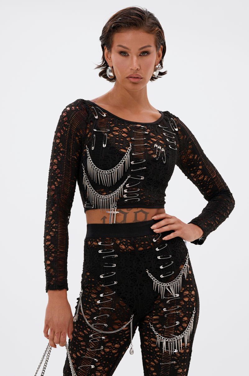 A woman stands confidently against a white background wearing the TRY ME LACE LONG SLEEVE WITH PINS, styled with matching pants featuring silver chains and safety pins for an edgy fashion look.