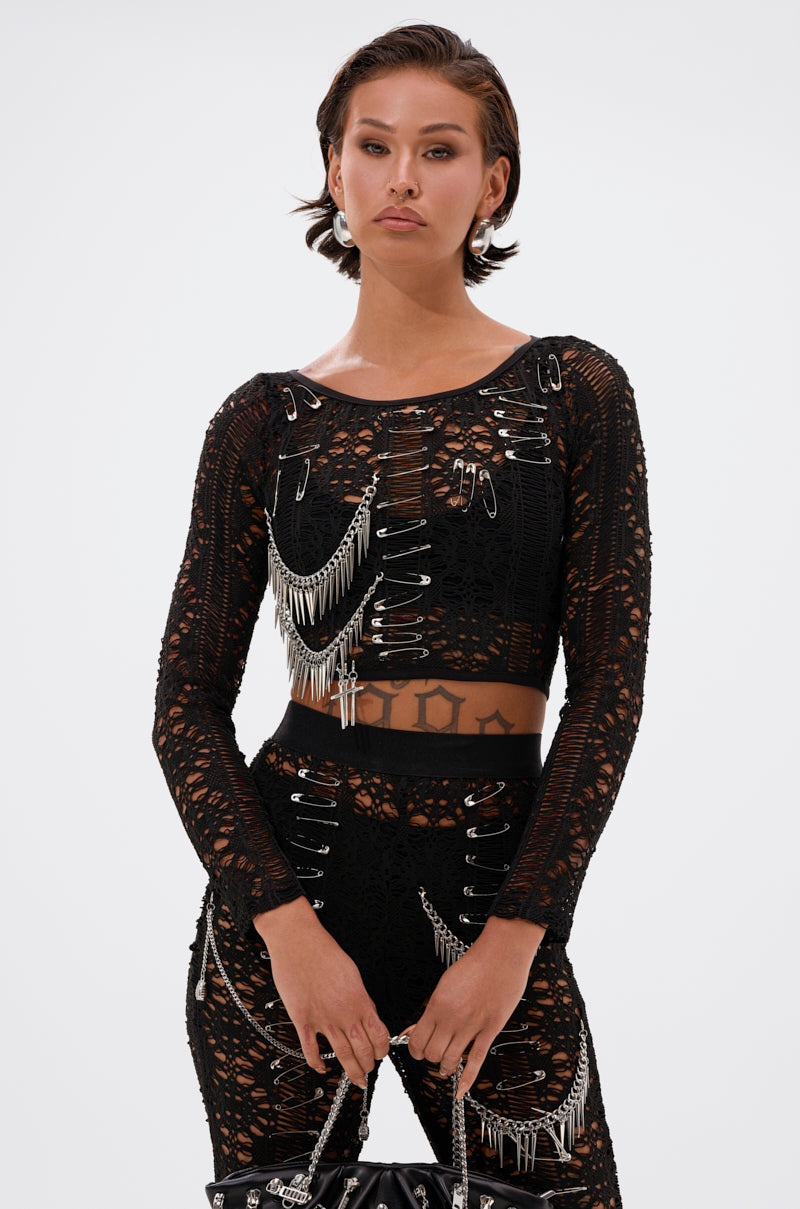 Wearing the TRY ME LACE LONG SLEEVE WITH PINS—black, sheer, long-sleeve, cropped, accented with large safety pins—a person pairs it with matching pants and a black handbag, posing against a plain light background.