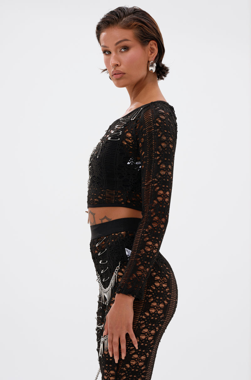 A woman with short brown hair models the TRY ME LACE LONG SLEEVE WITH PINS, an edgy cropped lace top, styled with a high-waisted skirt. She stands in profile against a white background, gazing confidently at the camera.
