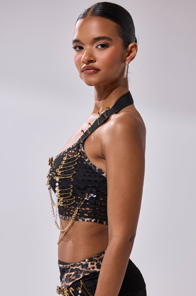A woman with slicked-back hair confidently poses in the FEELING EXTRA MESH TOP WITH BEADS, featuring gold safety pins, chains, and leopard print trim, paired with matching bottoms against a light gray background.