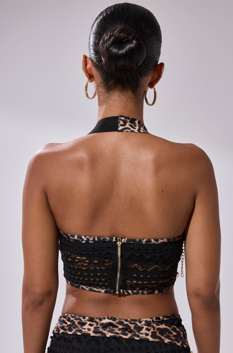 A woman with her hair in a bun, wearing gold hoop earrings and the FEELING EXTRA MESH TOP WITH BEADS, featuring a black lace mesh, zipper front, and leopard-print halter strap and waistband, is seen from the back.