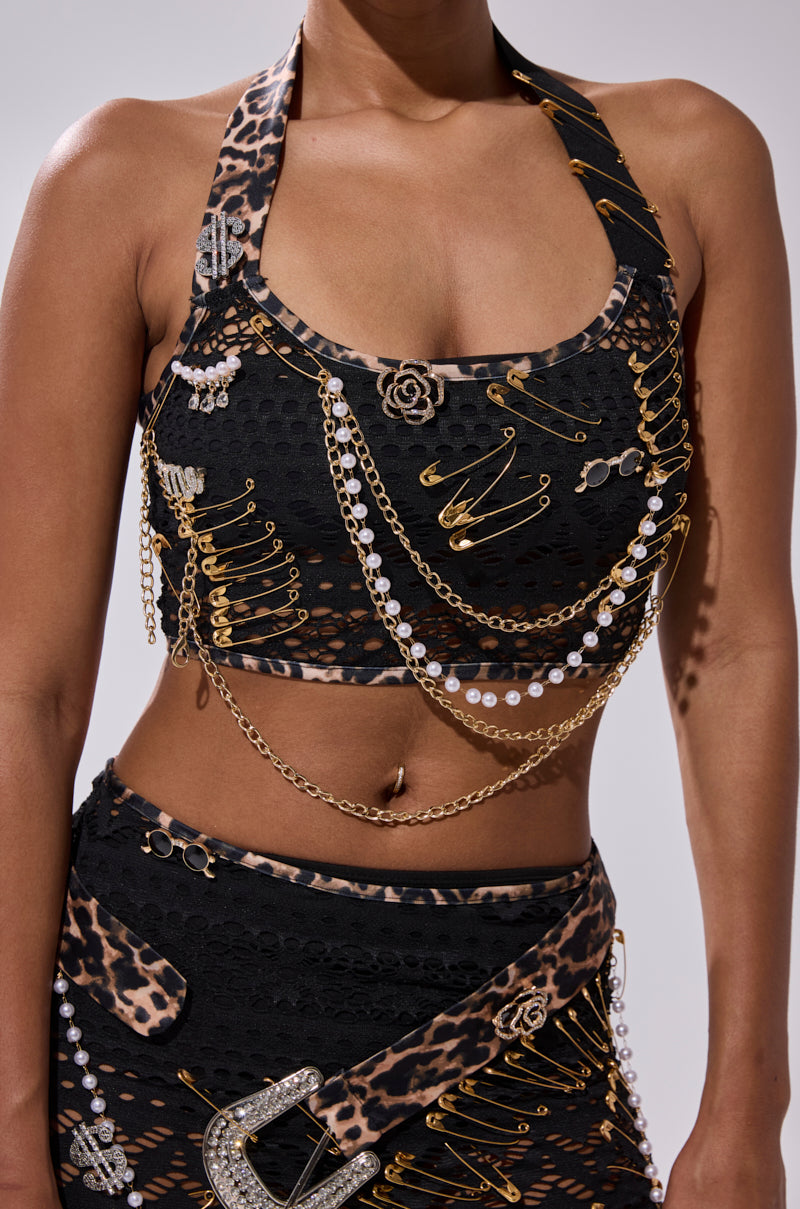 A woman wears the FEELING EXTRA MESH TOP WITH BEADS—a black crop top with pearl details—paired with a skirt featuring leopard print accents, gold safety pins, and chains. Only her torso is visible against a plain background.