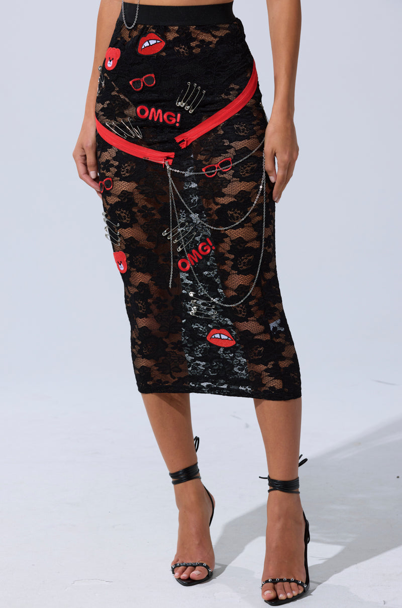 Wearing the OMG LACE MAXI SKIRT WITH EMBELLISHMENTS, featuring black floral lace, red and white lip, “OMG!”, glasses, and hand patches, plus silver chains. The look is completed with black high-heeled sandals.