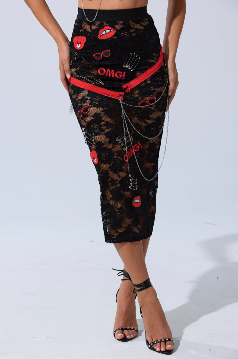 Wearing the OMG LACE MAXI SKIRT WITH EMBELLISHMENTS—black lace with red and white patches, "OMG!" text, lips, sunglasses, red zippers, and silver chains—paired with black strappy heels.
