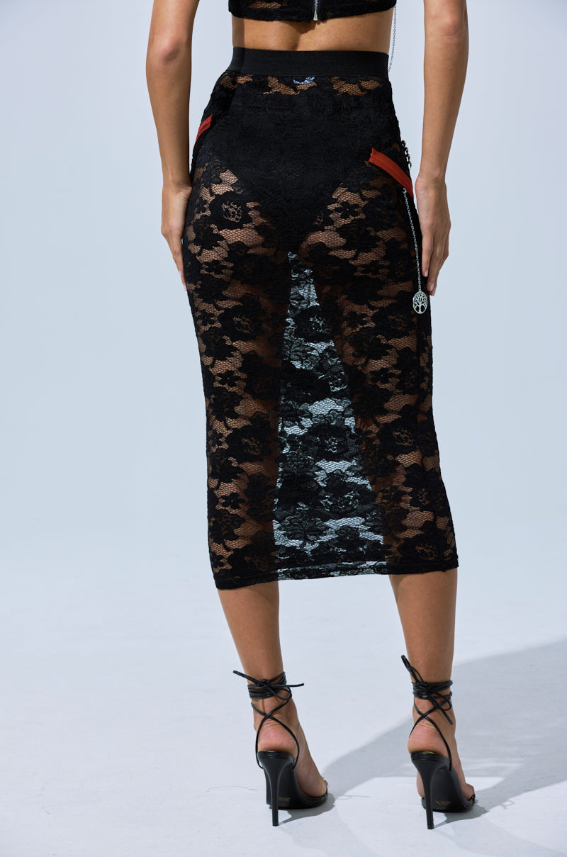 A person wears the OMG LACE MAXI SKIRT WITH EMBELLISHMENTS, featuring black floral lace with red accents, styled with high-heeled strappy sandals and photographed from the back against a plain light background.