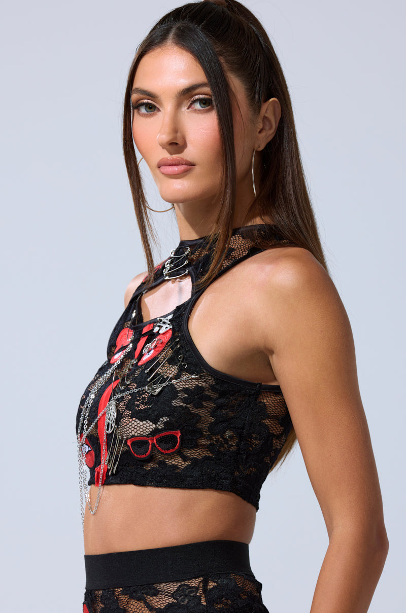 A woman with long brown hair in a high ponytail wears the OMG LACE TANK WITH EMBELLISHMENTS, featuring black lace and red and white decorative patches. She poses slightly sideways against a plain light background.