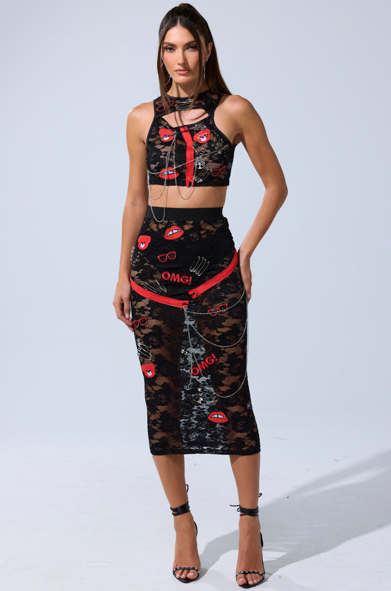 A woman faces forward in the OMG LACE TANK WITH EMBELLISHMENTS—a black lace crop top with red and white "OMG!" and eye patches—paired with a matching high-waisted skirt, black strappy heels, and silver accessories.