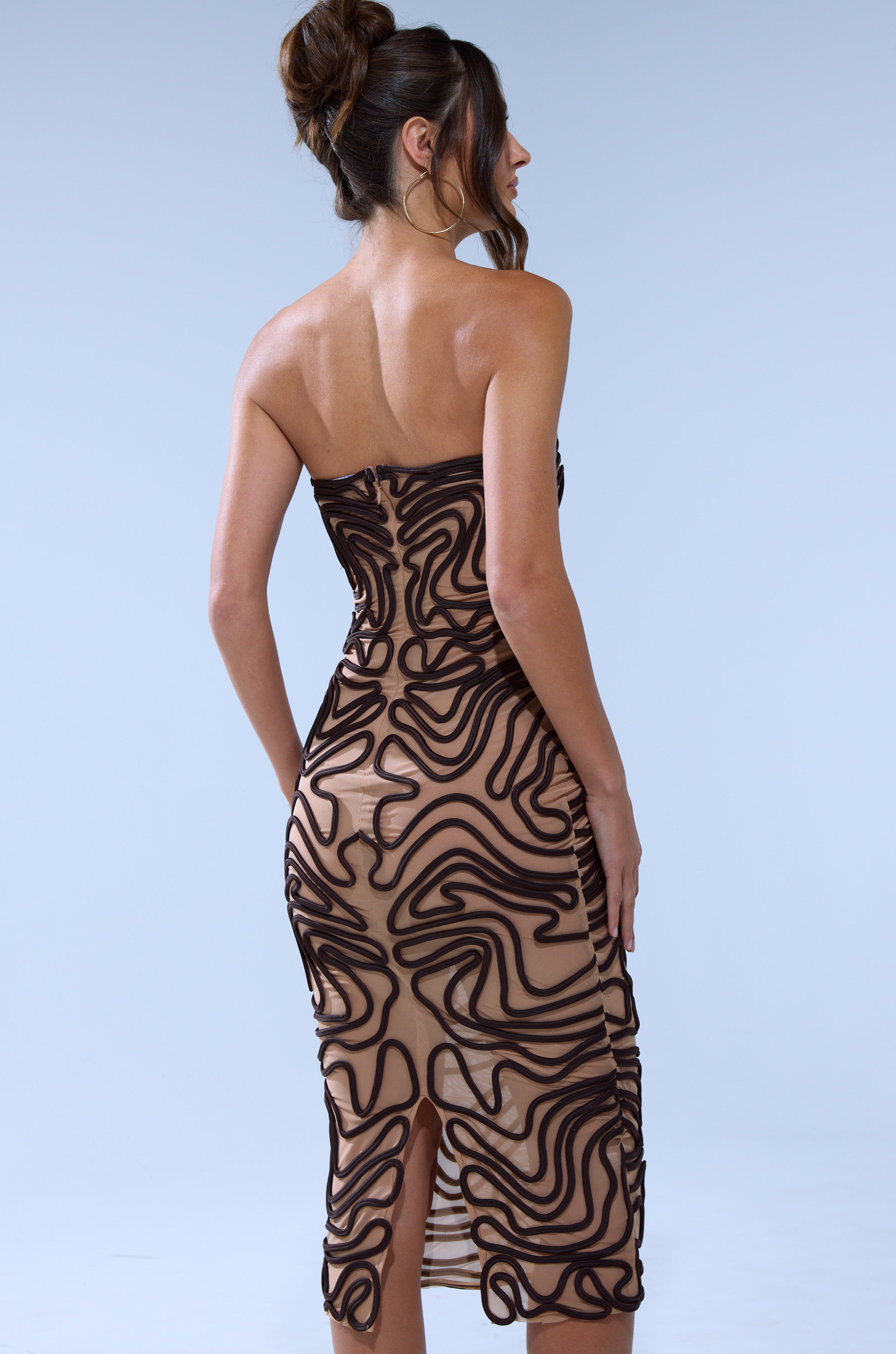 A woman stands facing away in the OUT OF THIS WORLD MIDI DRESS, a nude mesh midi with black abstract swirls and ribbed detailing, strapless neckline, against a plain light background.