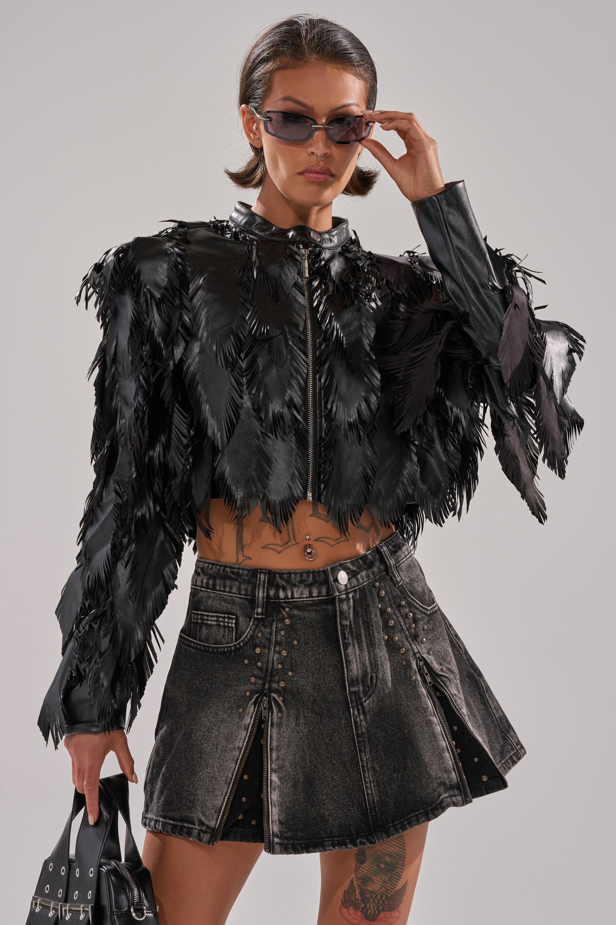 A woman in sunglasses wears the AVANT GARDE LASER CUT BOMBER—a black, feather-textured cropped jacket—with a high-waisted pleated denim mini skirt and studs. She carries a black handbag, showing her torso tattoos and striking a confident pose. 1