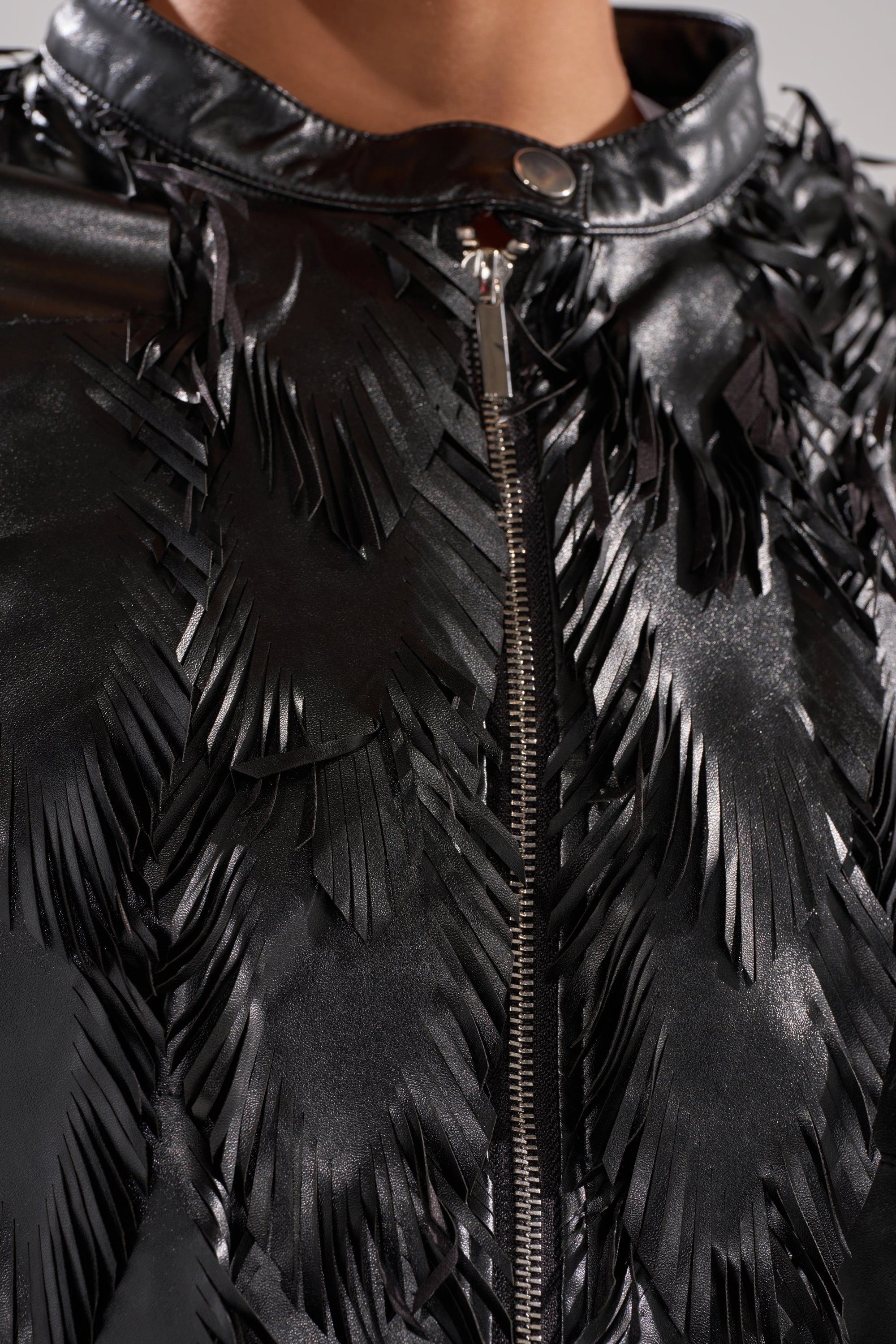 Close-up of the AVANT GARDE LASER CUT BOMBER in shiny black faux leather, featuring a zipper, high collar, and chevron-shaped layered fringe for a textured, dimensional look. 9
