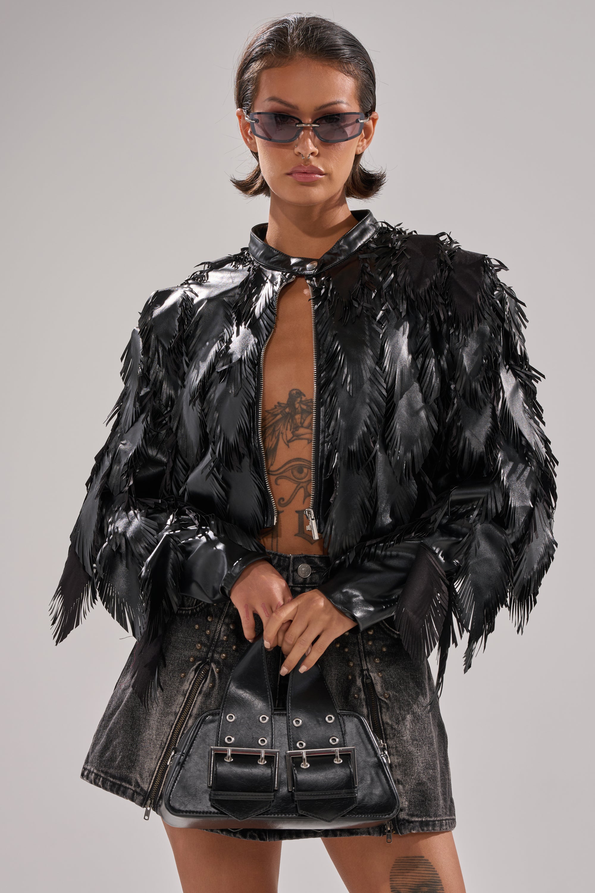 A woman in the AVANT GARDE LASER CUT BOMBER, sunglasses, and a short dark denim skirt stands holding a black handbag. She displays visible torso tattoos and exudes confidence. 0