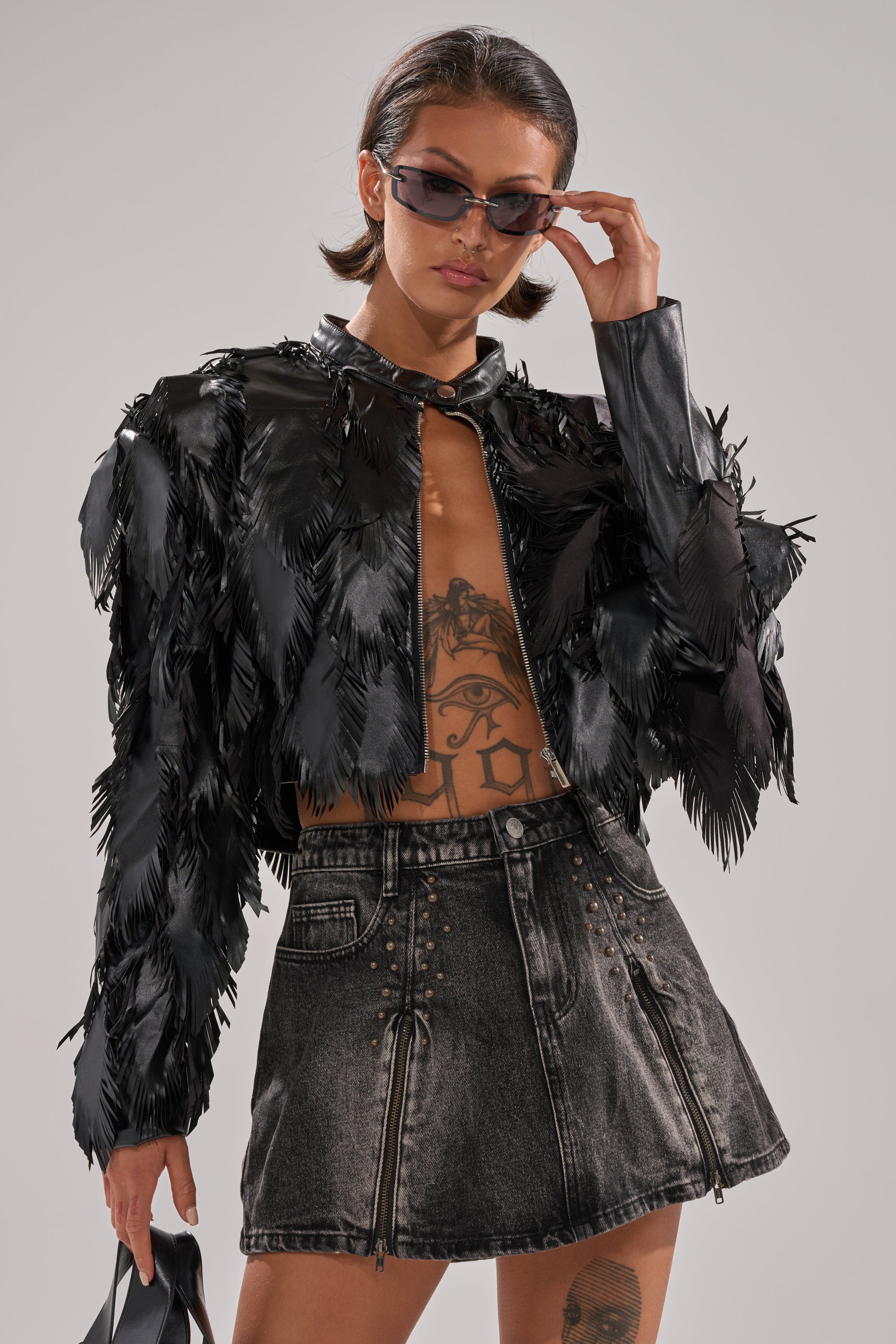 Wearing dark sunglasses and the AVANT GARDE LASER CUT BOMBER over a high-waisted black denim mini skirt, they stand confidently with their open jacket showing a large eye tattoo on their torso. 2
