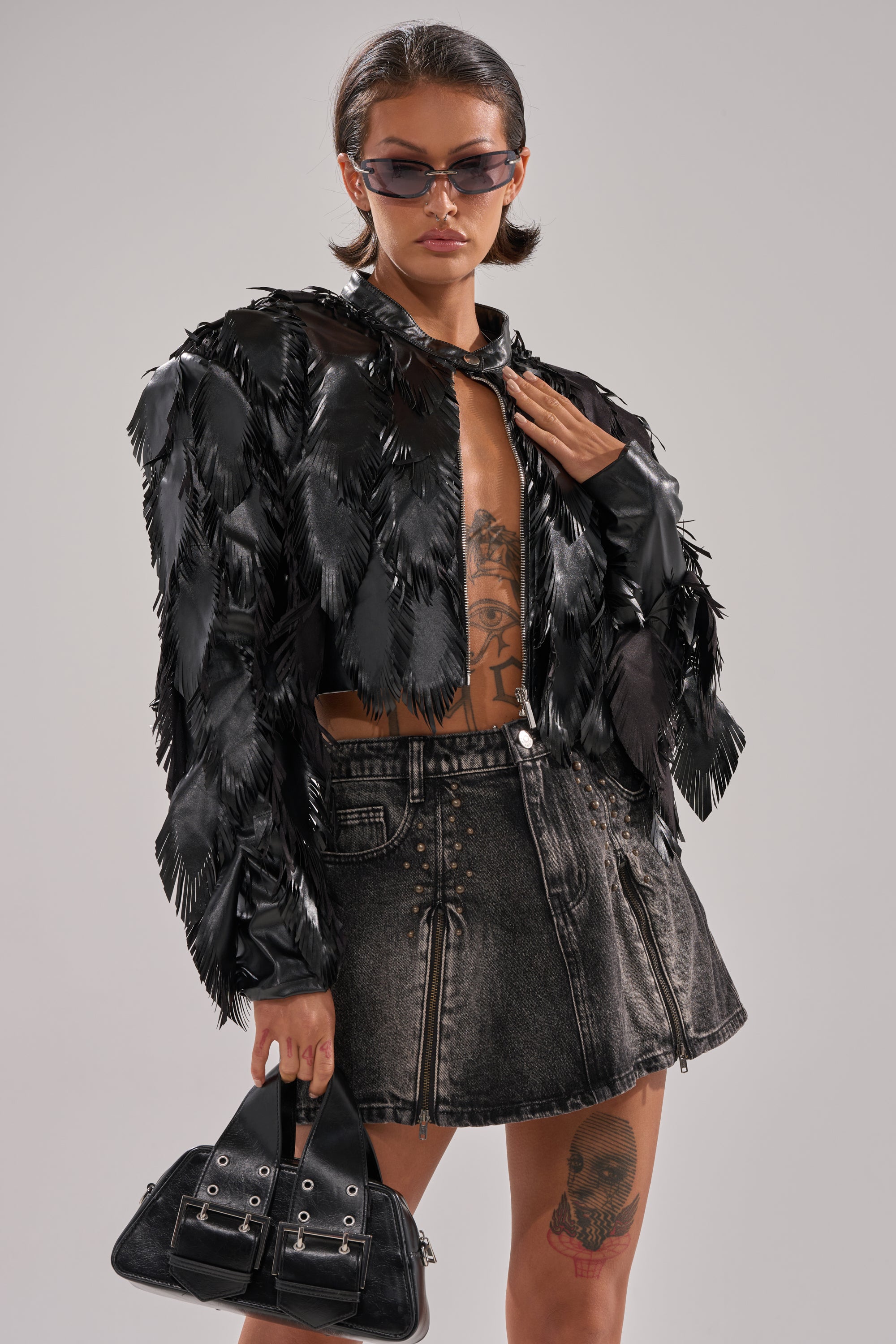 A woman in sunglasses poses confidently against a plain background, wearing the AVANT GARDE LASER CUT BOMBER over an open shirt with chest tattoos, a faded black denim mini skirt, and holding a black handbag. 3