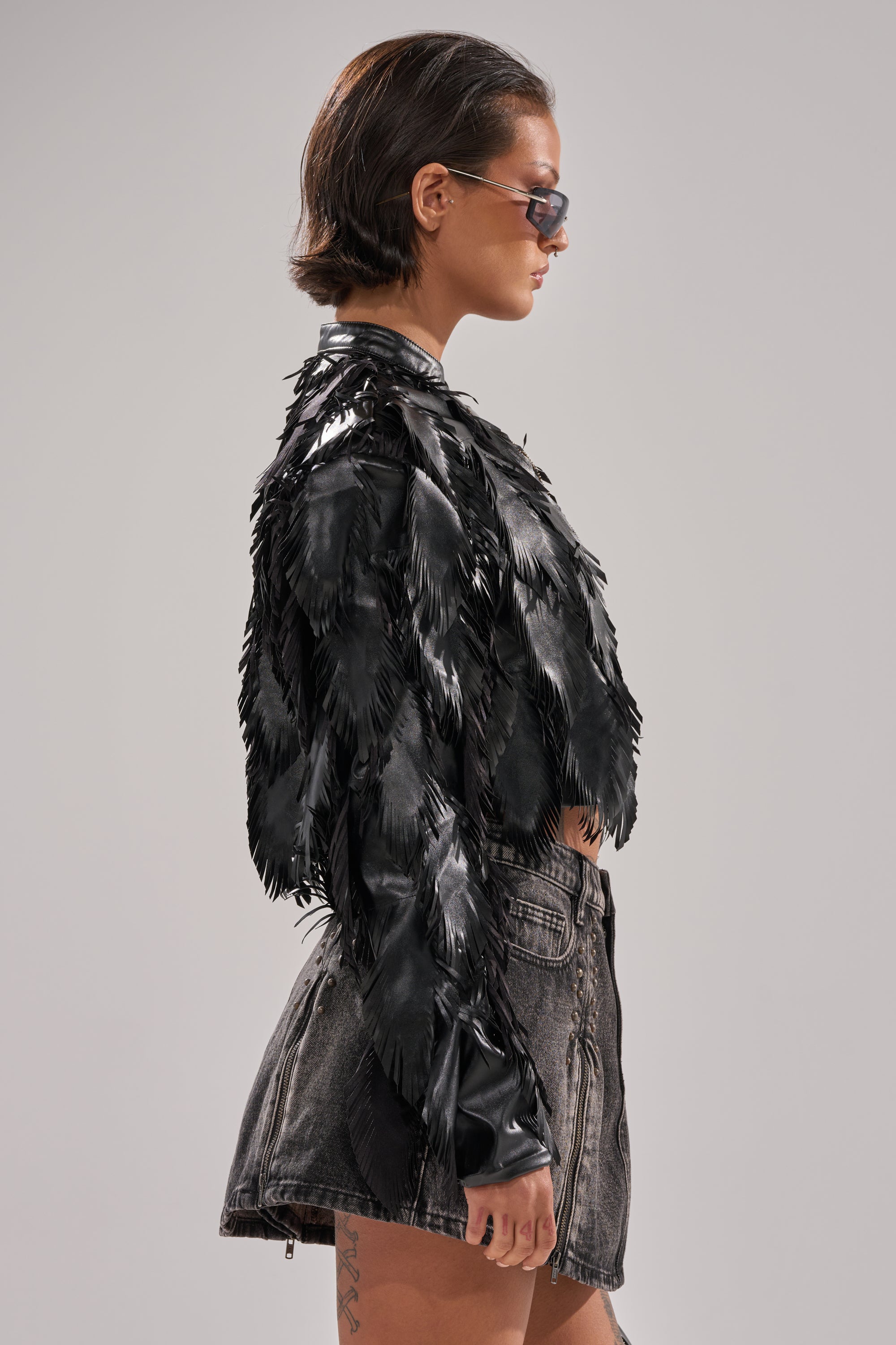 A person with short dark hair and sunglasses models the AVANT GARDE LASER CUT BOMBER, featuring feather-like details, paired with a high-waisted, faded black denim skirt. They stand sideways against a plain background. 4