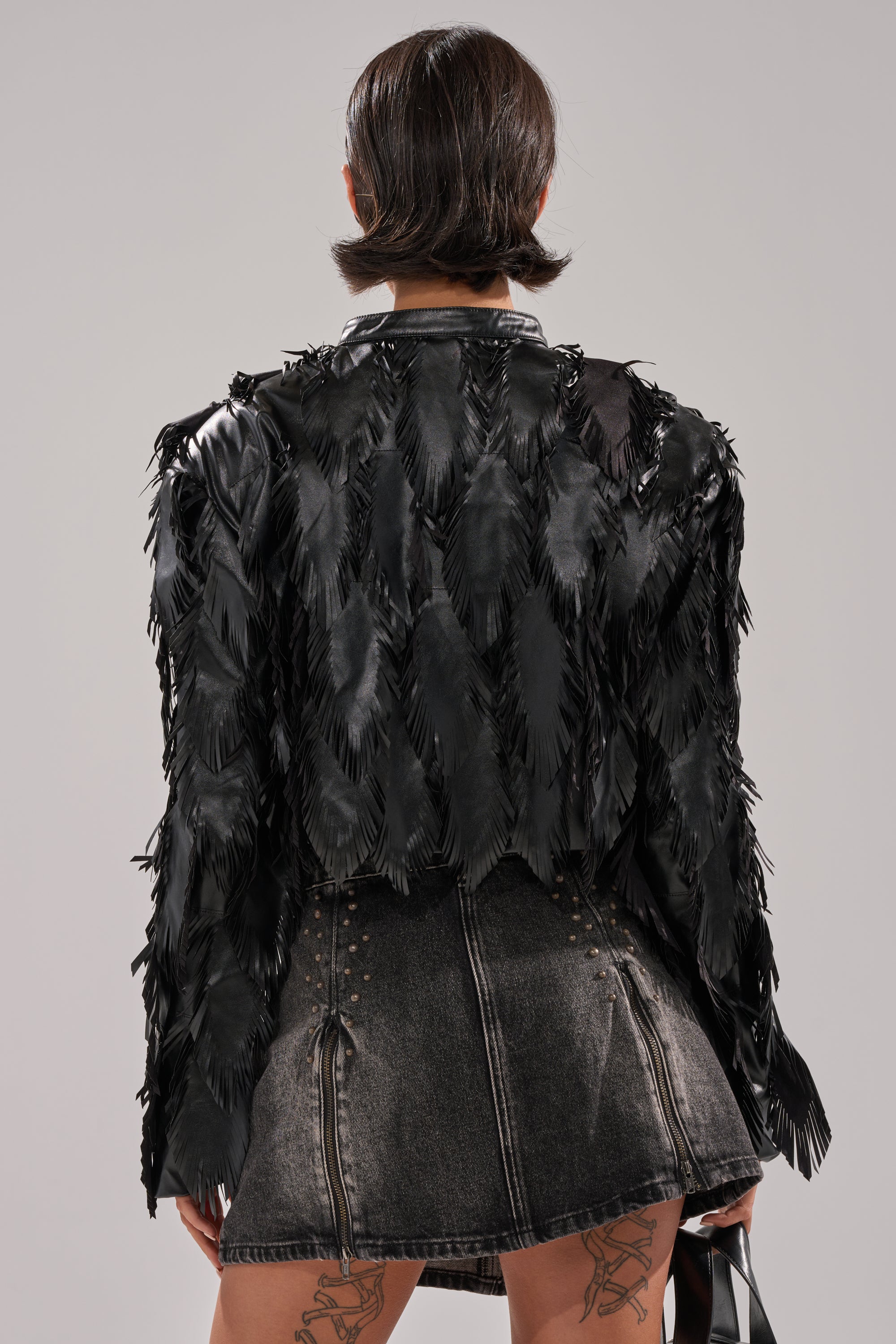 A person with short dark hair, seen from behind, wears the AVANT GARDE LASER CUT BOMBER and a faded gray denim mini skirt. Tattoo designs are visible on their thighs, and they hold a black bag in their right hand. 5