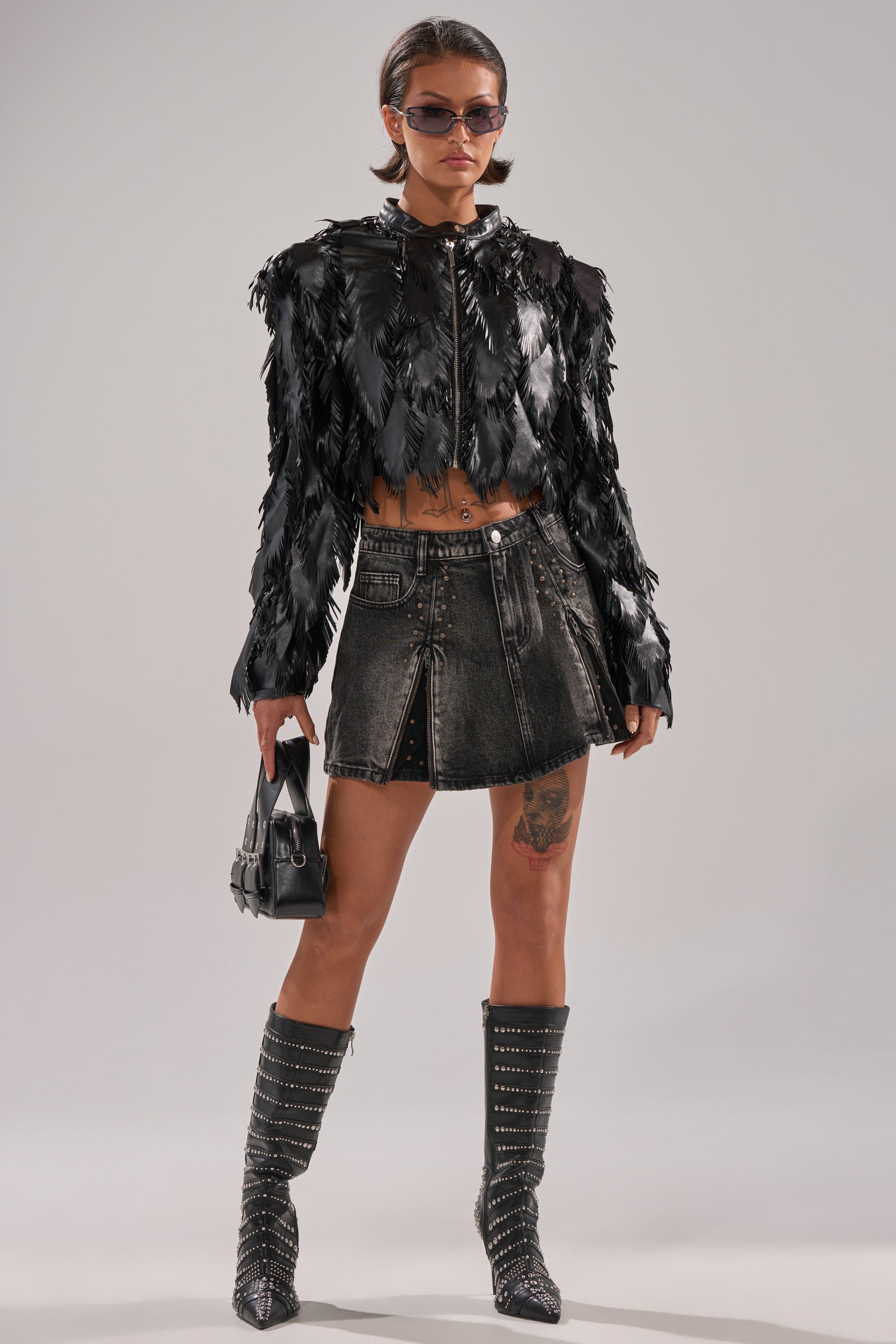A person stands confidently in sunglasses, the AVANT GARDE LASER CUT BOMBER, a short black denim skirt, studded knee-high boots, holding a small black handbag and showing a thigh tattoo. 8