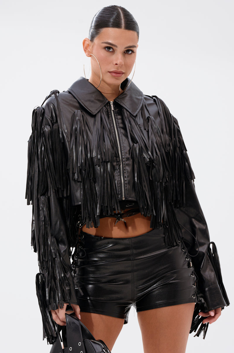 A woman stands confidently in the SIERRA TASSEL MOTO— a black cropped faux leather fringe jacket with matching high-waisted shorts and bag. Her slicked-back hair, hoop earrings, and poised expression complete the bold look.
