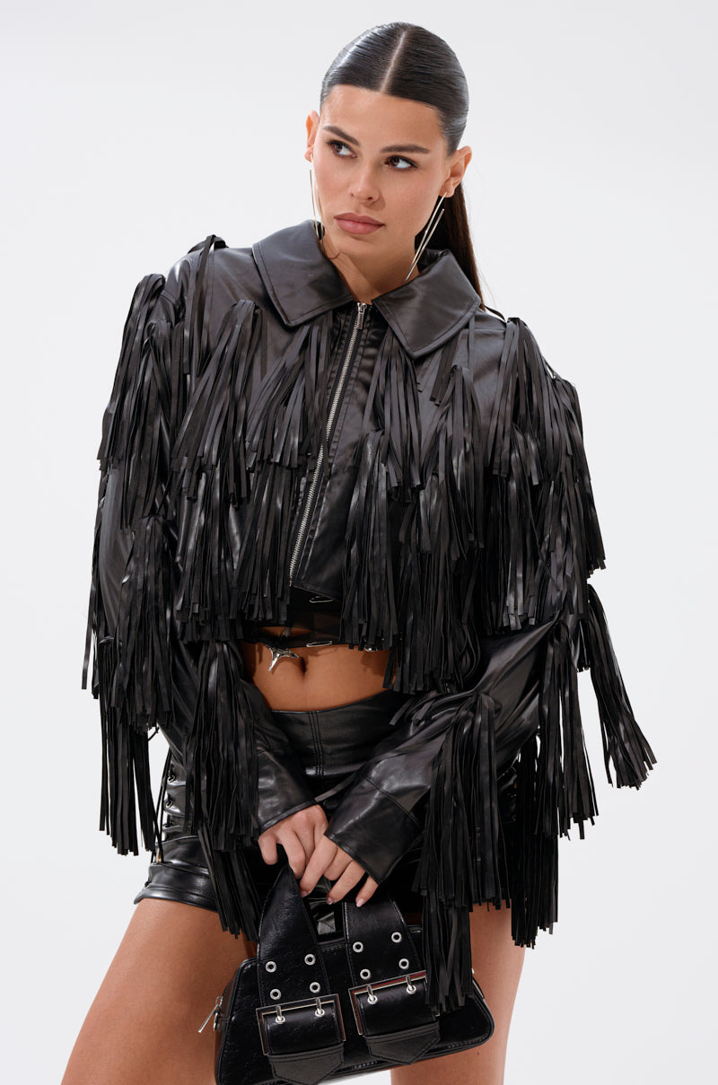A woman with straight dark hair in a sleek ponytail wears the SIERRA TASSEL MOTO faux leather fringe jacket with matching black leather shorts and holds a small black purse against a plain white background.