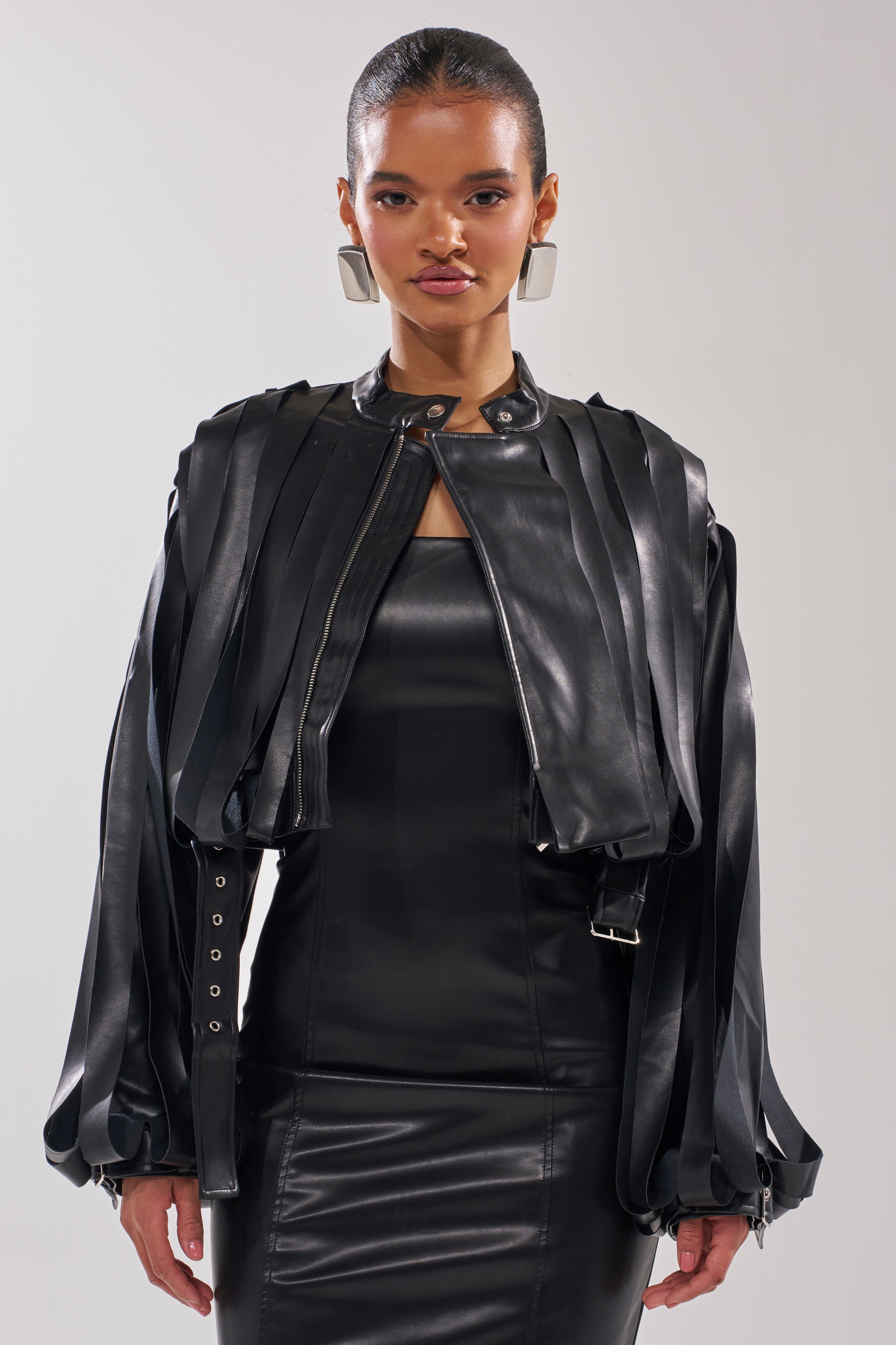 A woman with slicked-back hair wears the GONE WEST STRAPPY MOTO—a shiny black faux leather jacket with vertical strips and wide sleeves—posing against a plain light background.