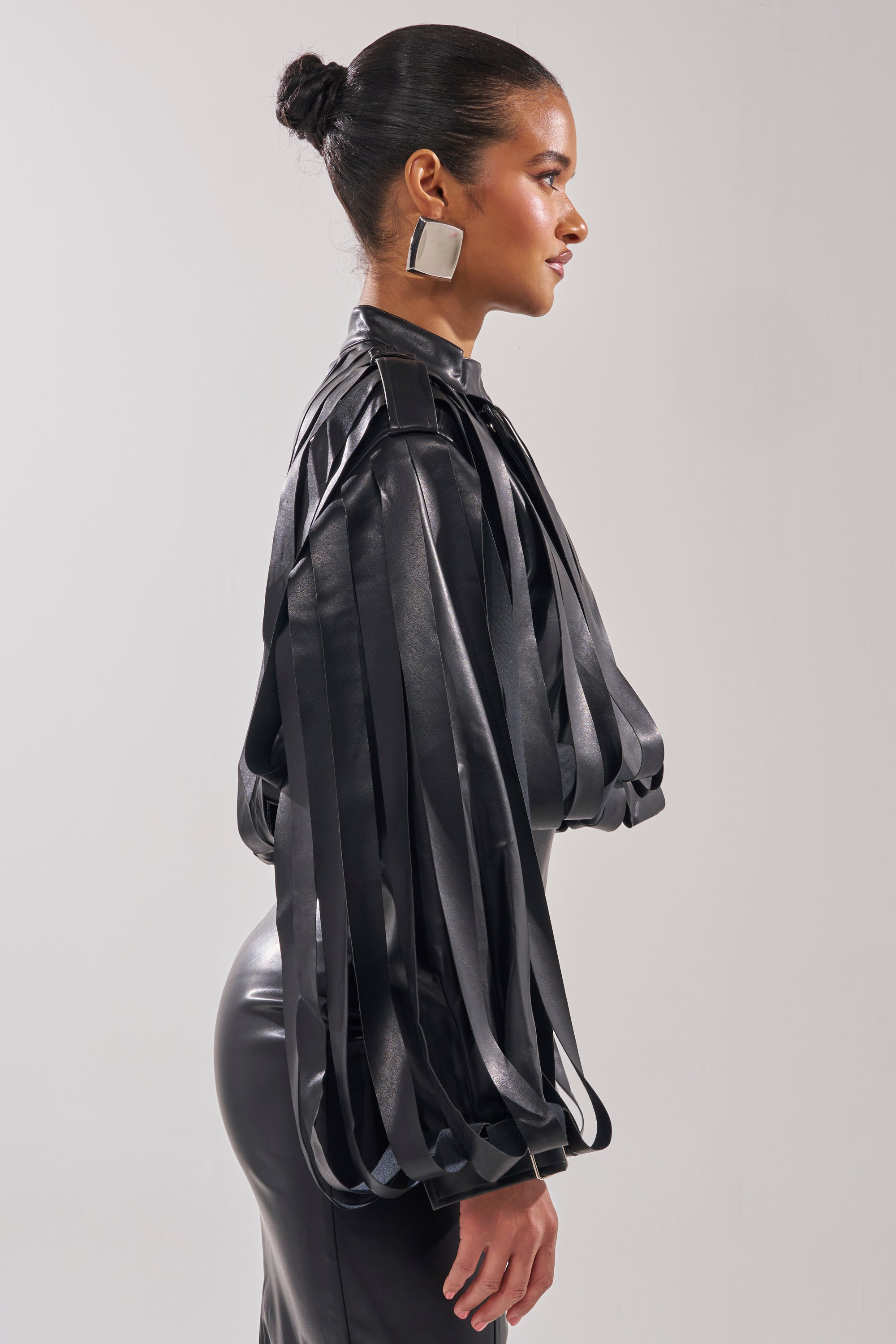 A woman in profile poses against a plain background, wearing the GONE WEST STRAPPY MOTO—a black, shiny faux leather blouse with long strips and voluminous sleeves. Her hair is styled in a sleek bun with bold square metallic earrings.