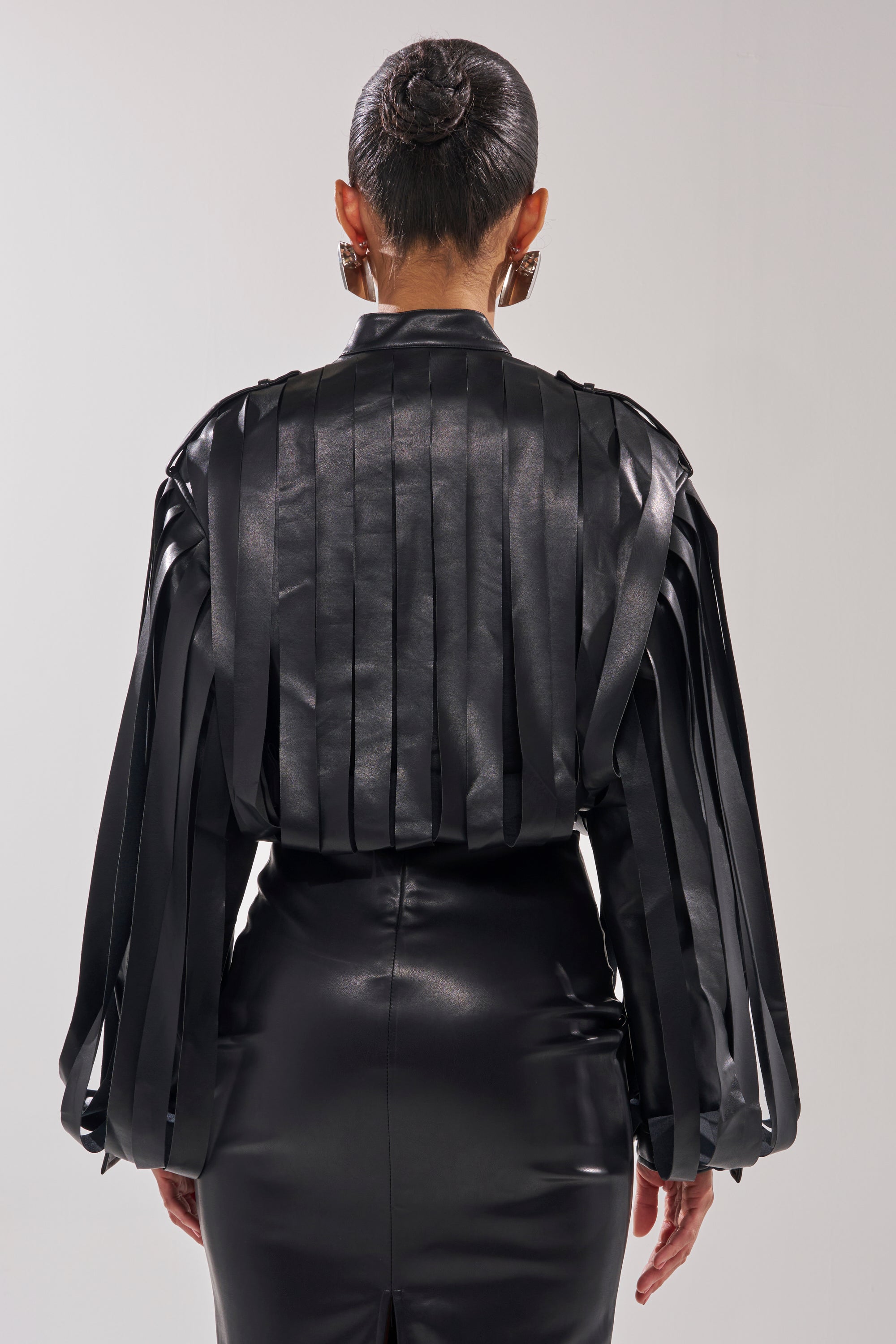 A person faces away, wearing the GONE WEST STRAPPY MOTO—a black, glossy faux leather long-sleeve top with vertical ribbon-like strips—paired with a matching skirt, hair in a neat bun, and large geometric earrings.