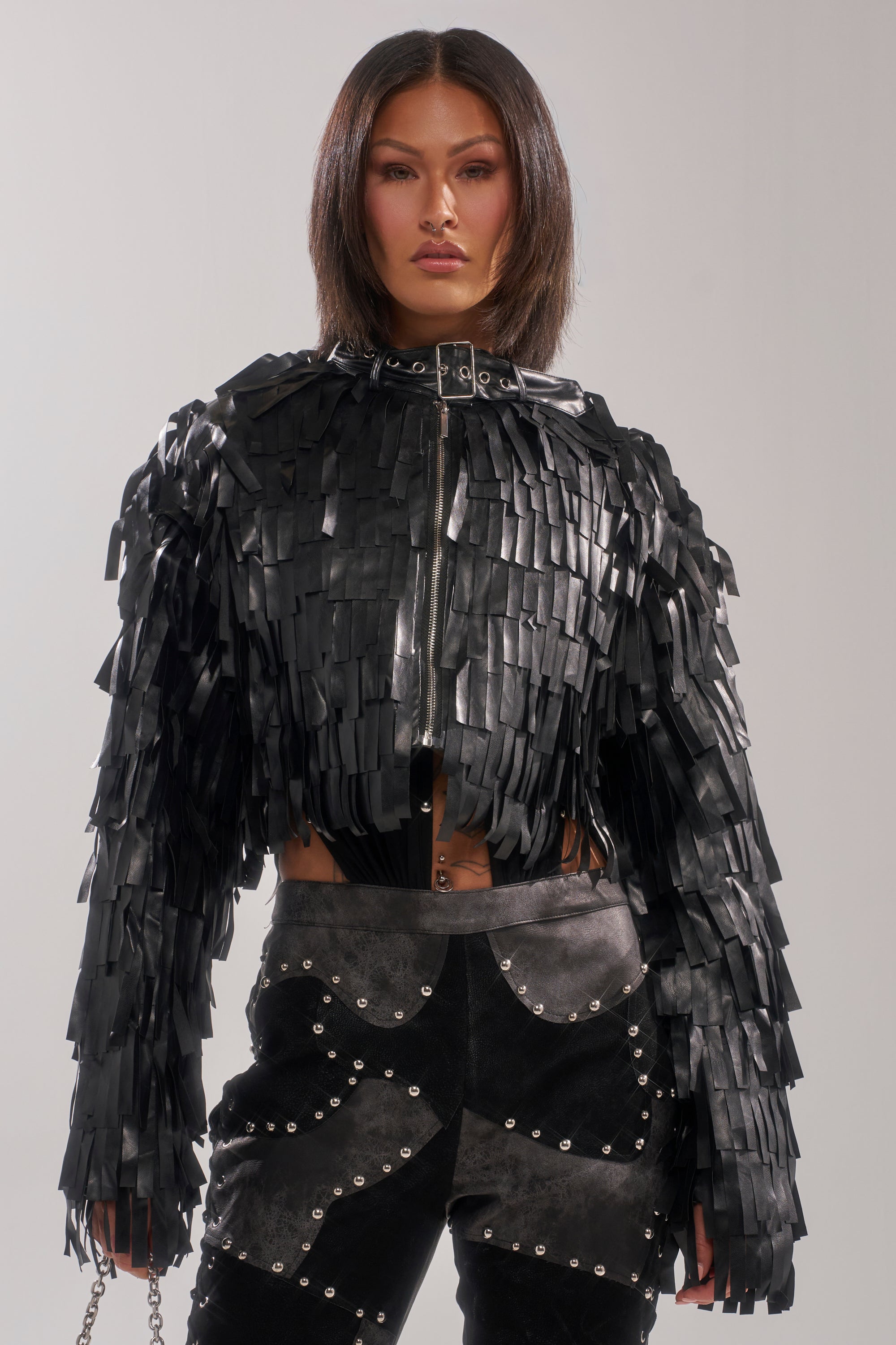 A person with shoulder-length dark hair wears the RIDE EM FRINGE MOTO, a shiny black jacket with layered fringe strips, paired with high-waisted black pants featuring silver studs. They stand against a plain, light background.